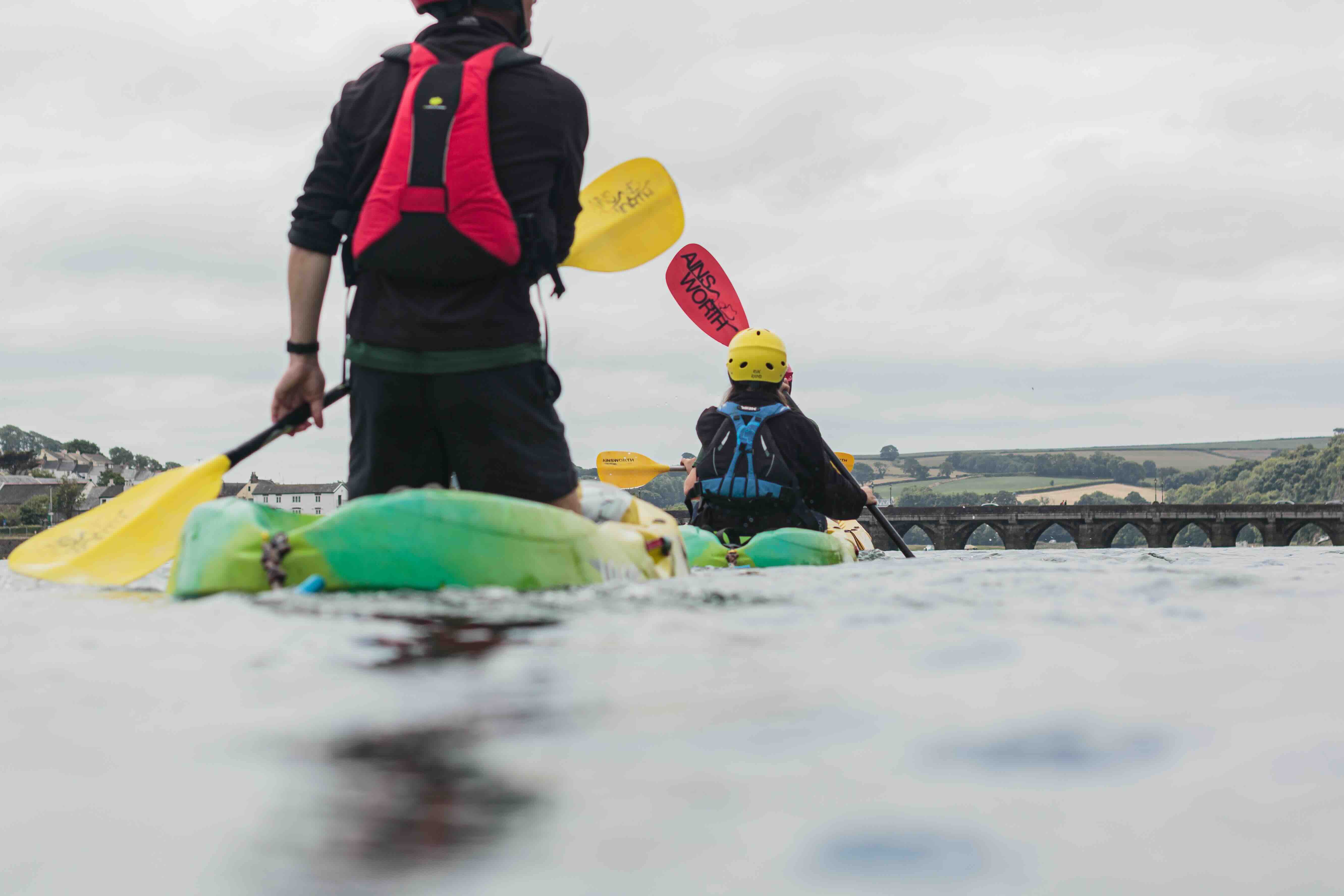 Two people kayaking on a river heading towards a stone bridge with arched openings under a cloudy sky.