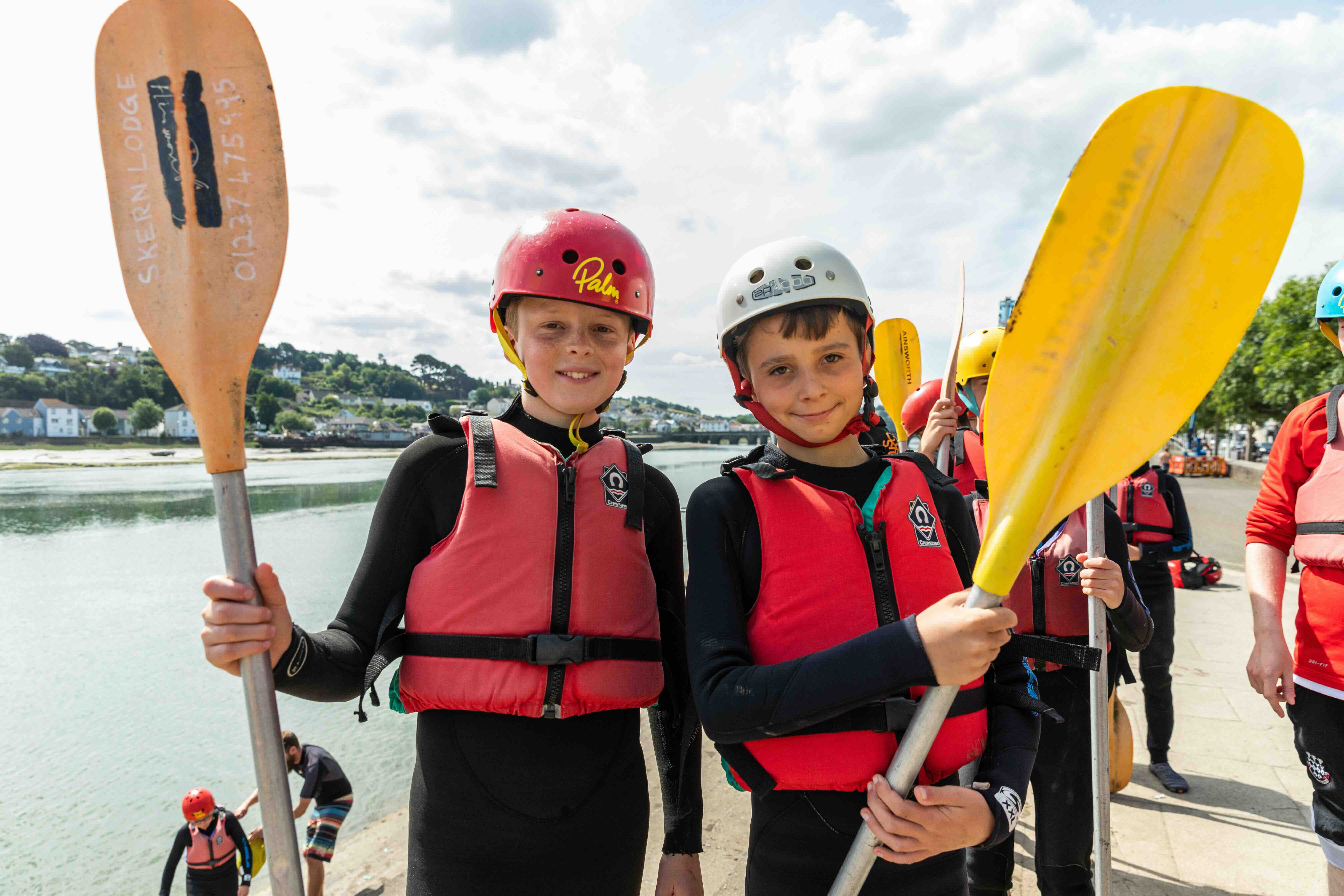 Two boys wearing red life jackets and helmets holding kayak paddles beside a river, with other kayakers and a town in the background.