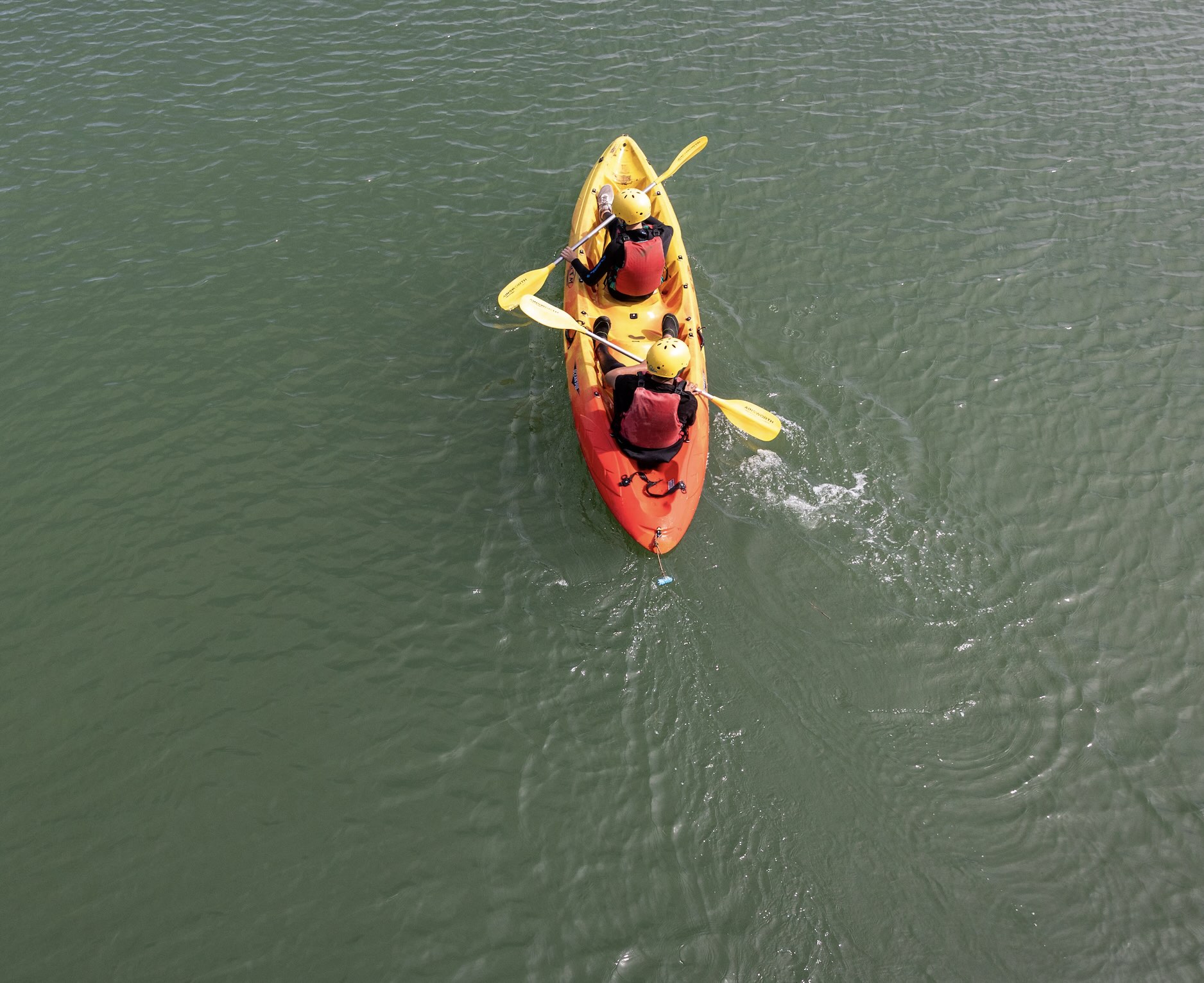 Two people wearing yellow helmets and red life jackets paddling a tandem kayak on green water.