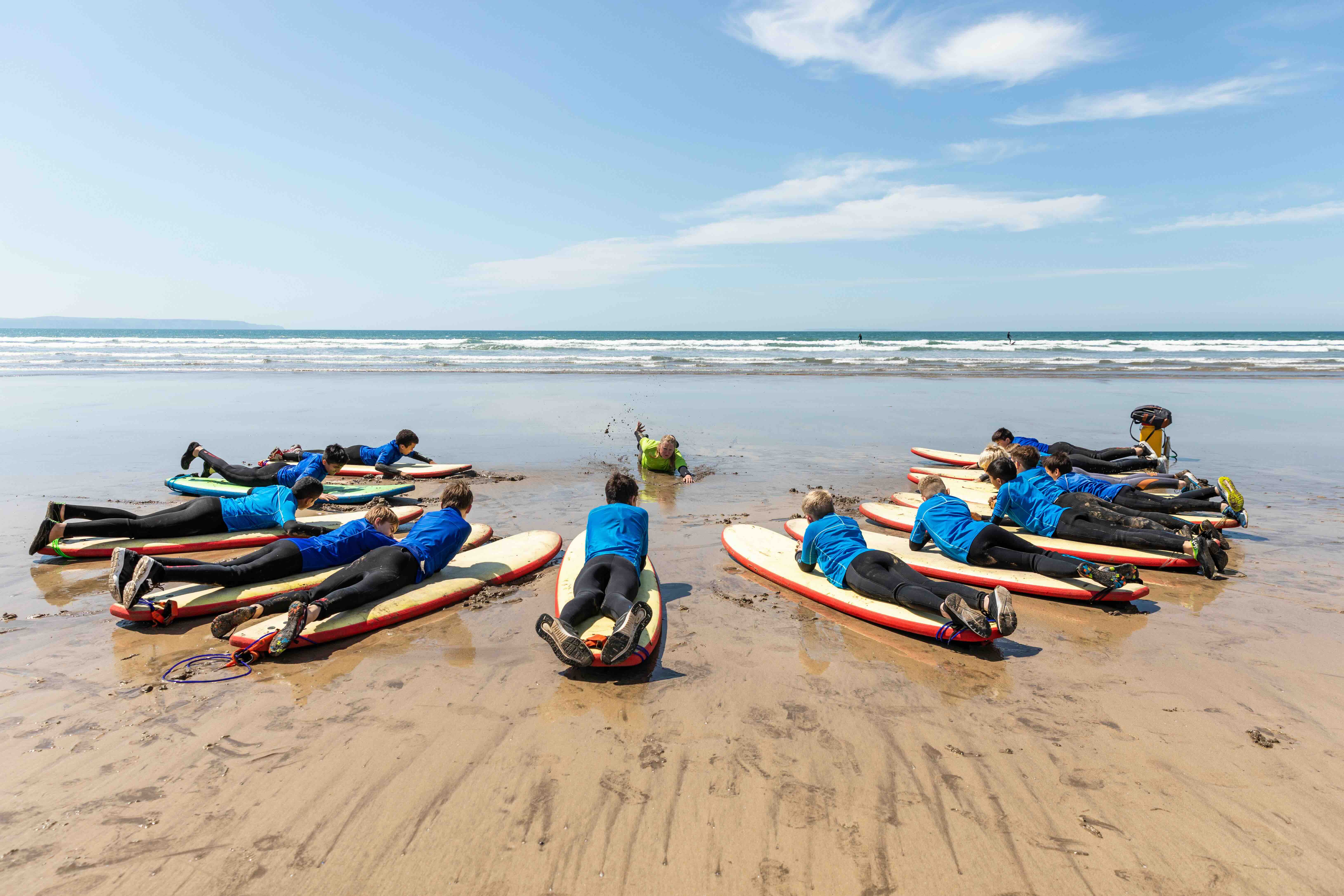 Group of children lying on surfboards on the sandy beach practicing paddling with instructor in the water on a sunny day.