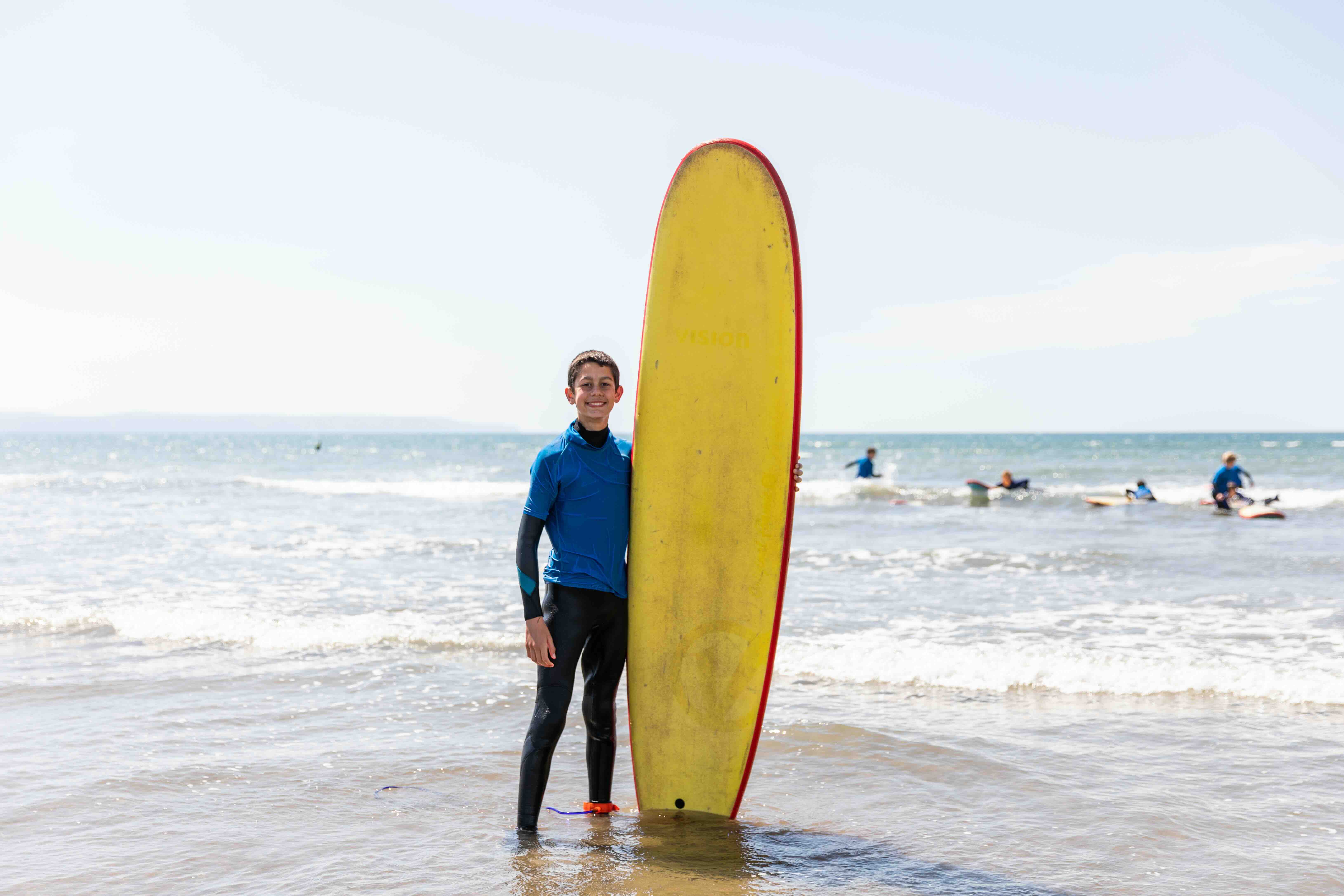 Smiling boy in wetsuit standing in shallow water holding a tall yellow surfboard with people surfing in the background.