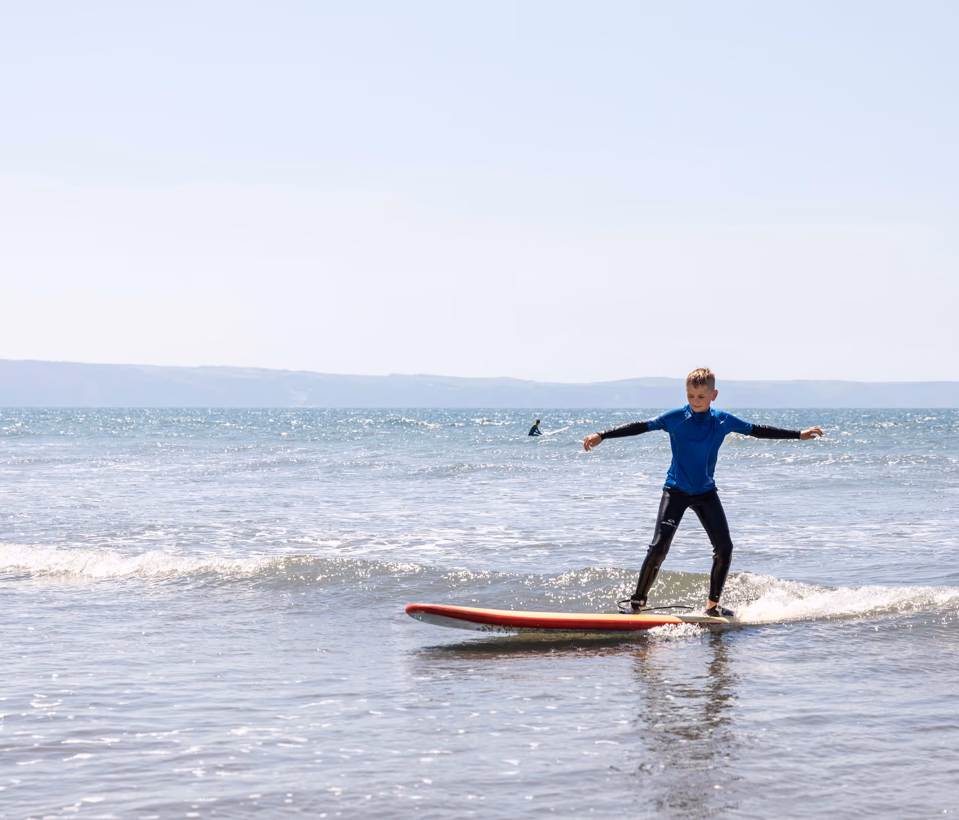 Young boy in wetsuit balancing on a red surfboard on a small wave at the beach.