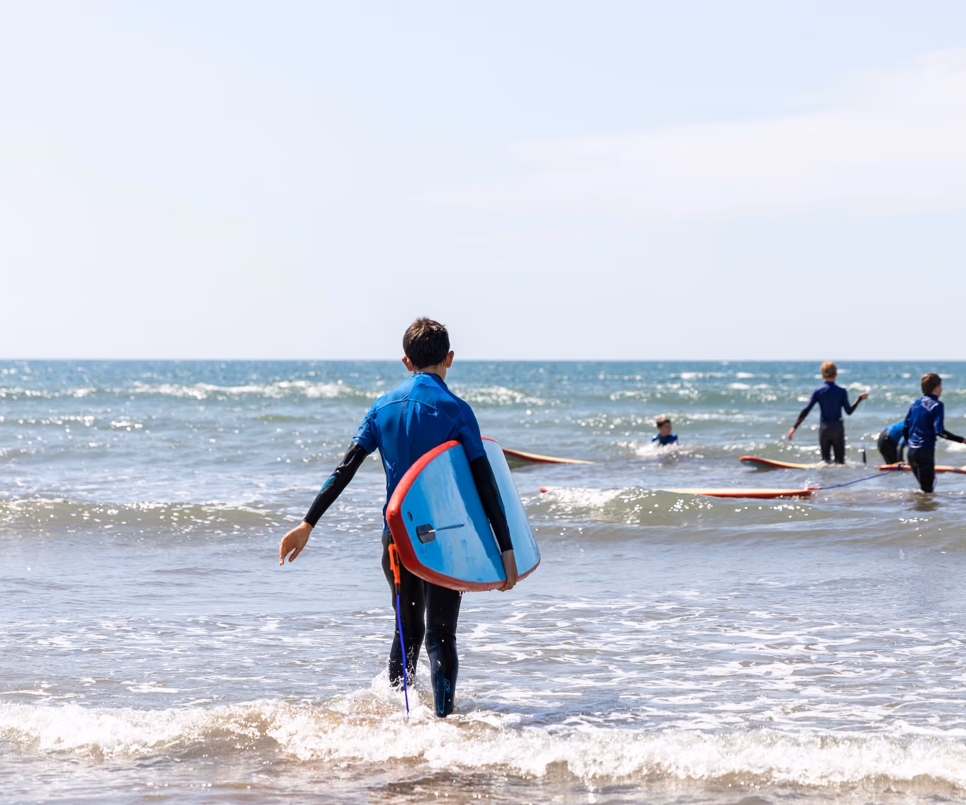 Person in a wetsuit carrying a surfboard walking into the ocean with others in wetsuits and surfboards in the water.