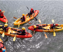 A group of people wearing helmets paddling in colorful kayaks on calm water.