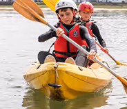 Two children wearing helmets and life jackets paddling a yellow kayak on calm water.