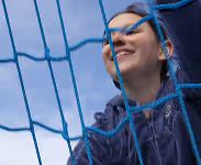 Smiling young woman behind a blue sports net wearing a navy hoodie against a cloudy sky.