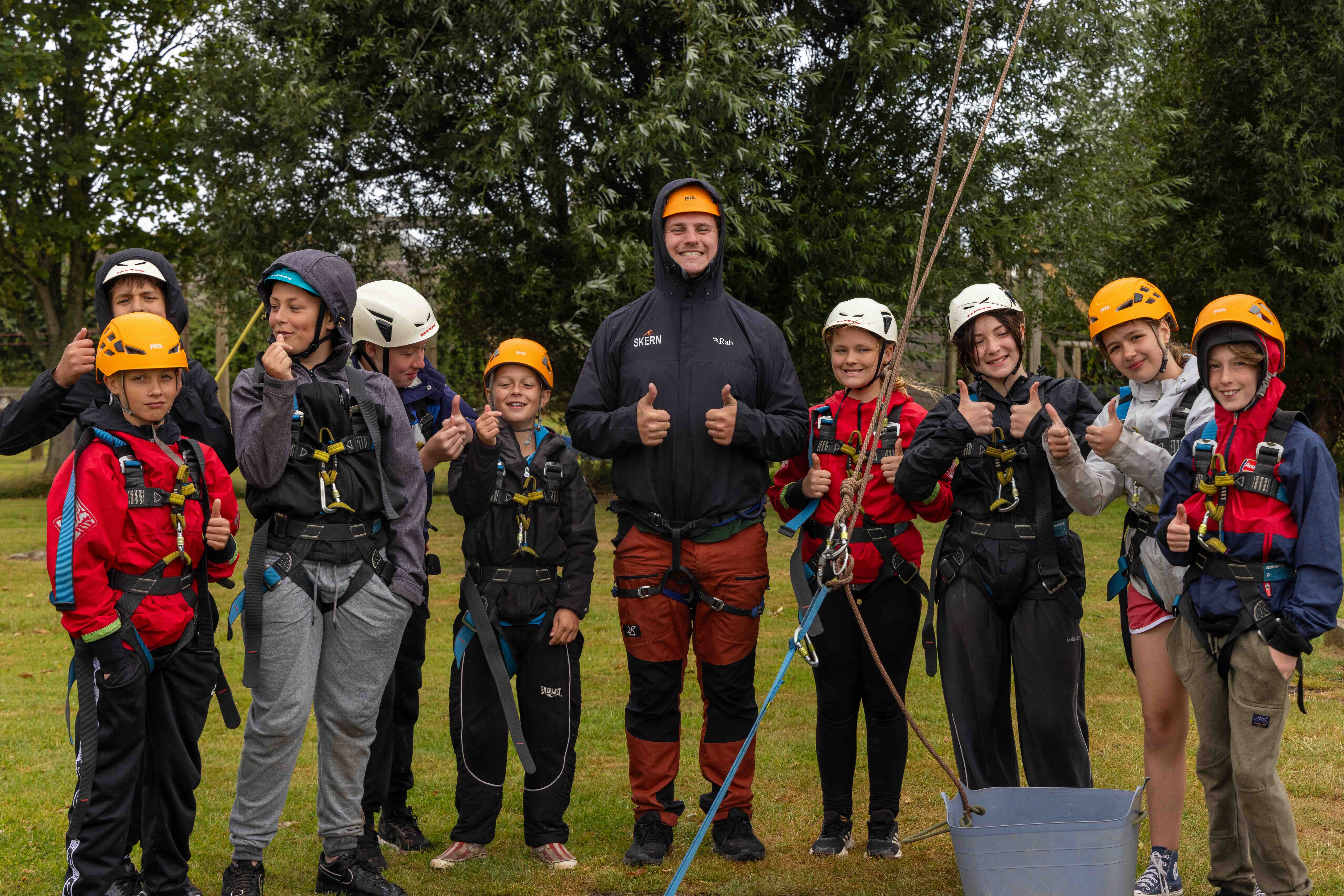Group of children and an instructor outdoors wearing climbing helmets and harnesses, all giving thumbs up.