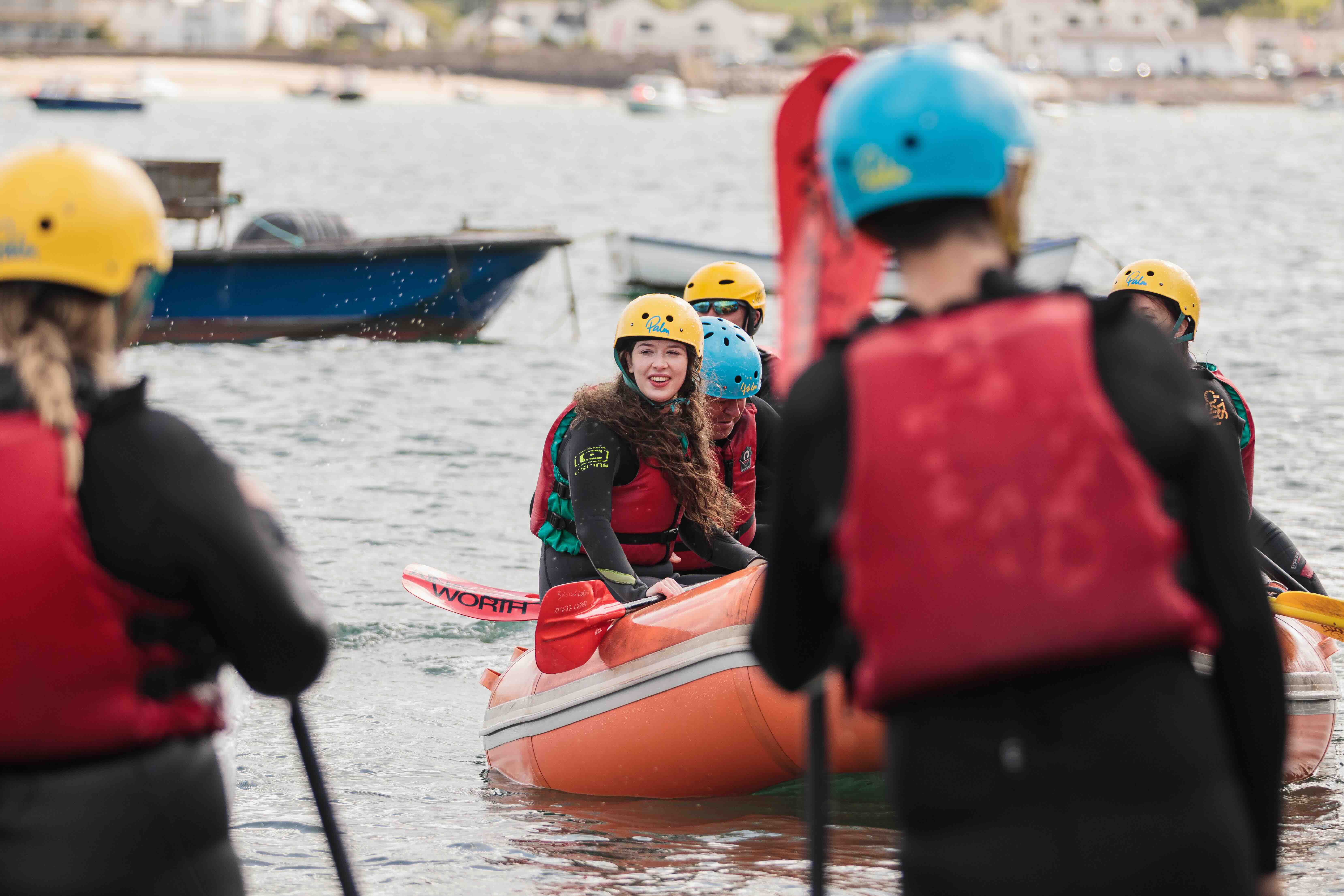 Group of people wearing helmets and life jackets, paddling an orange inflatable raft on water near a shore.