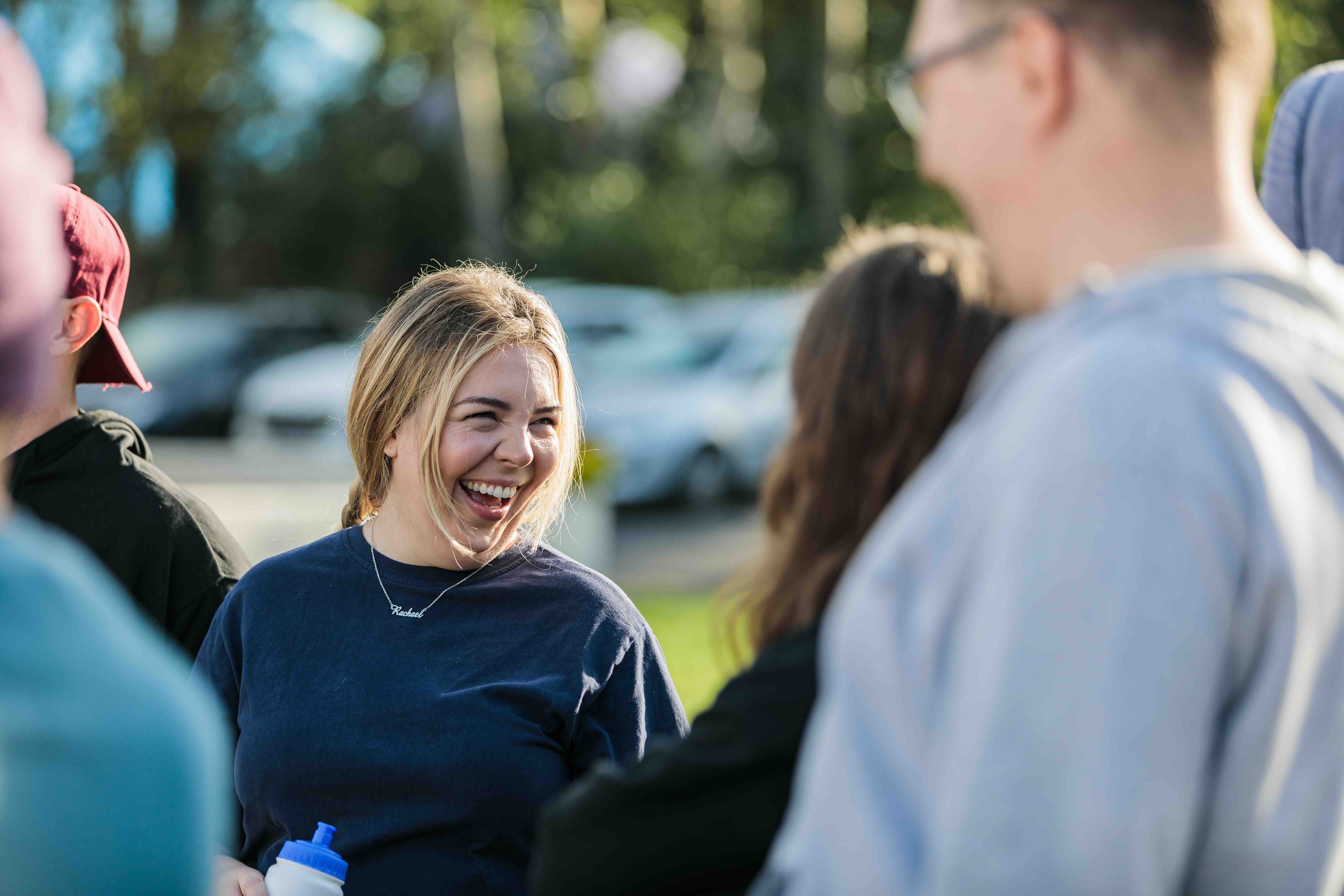 Smiling blonde woman in a dark shirt holding a water bottle, engaging with a group outdoors in daylight.