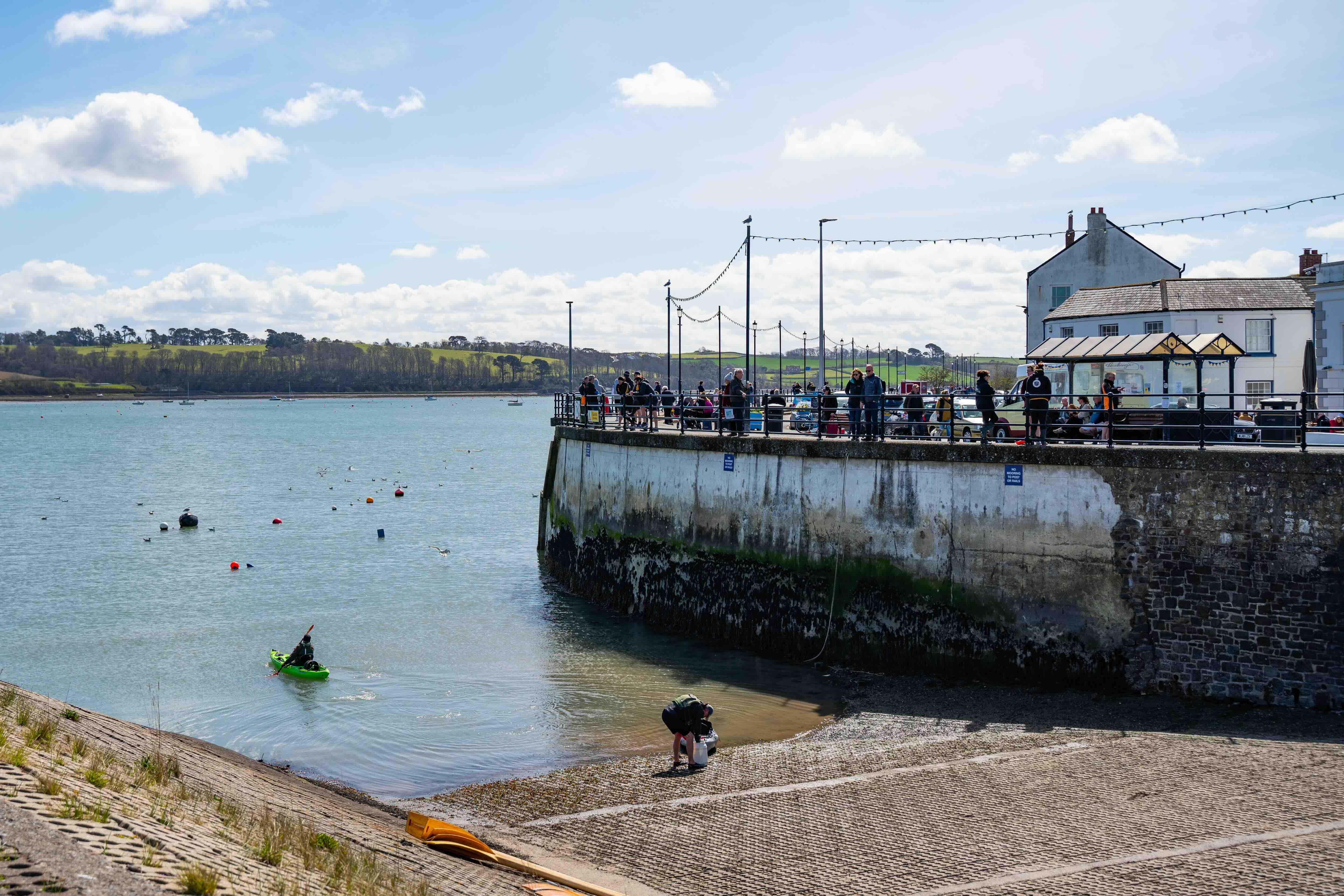 People gathered on a stone pier with buildings nearby overlooking a calm bay with a kayaker paddling in the water under a partly cloudy sky.