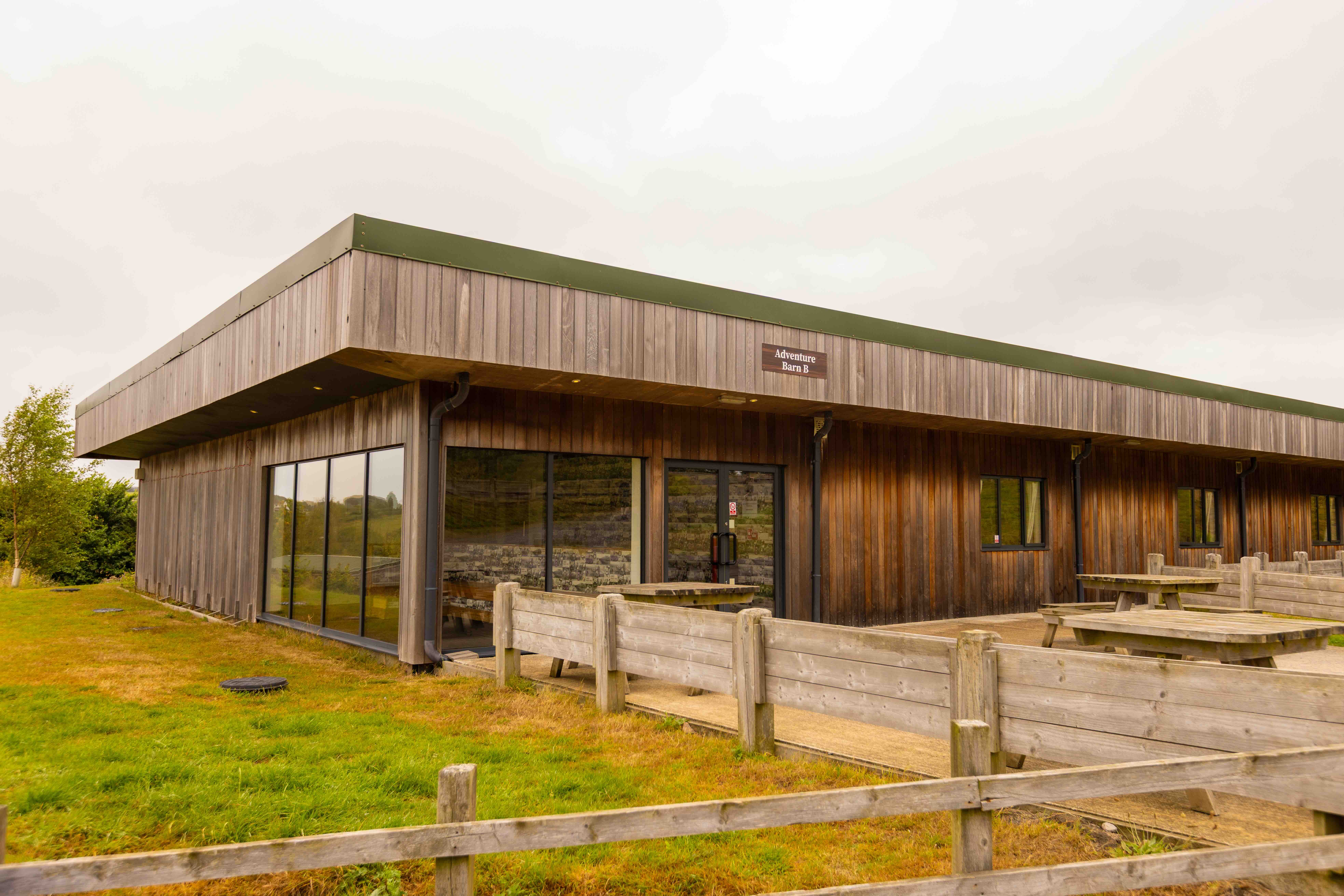 Wooden building named Adventure Barn B with outdoor picnic tables and fenced grassy area.
