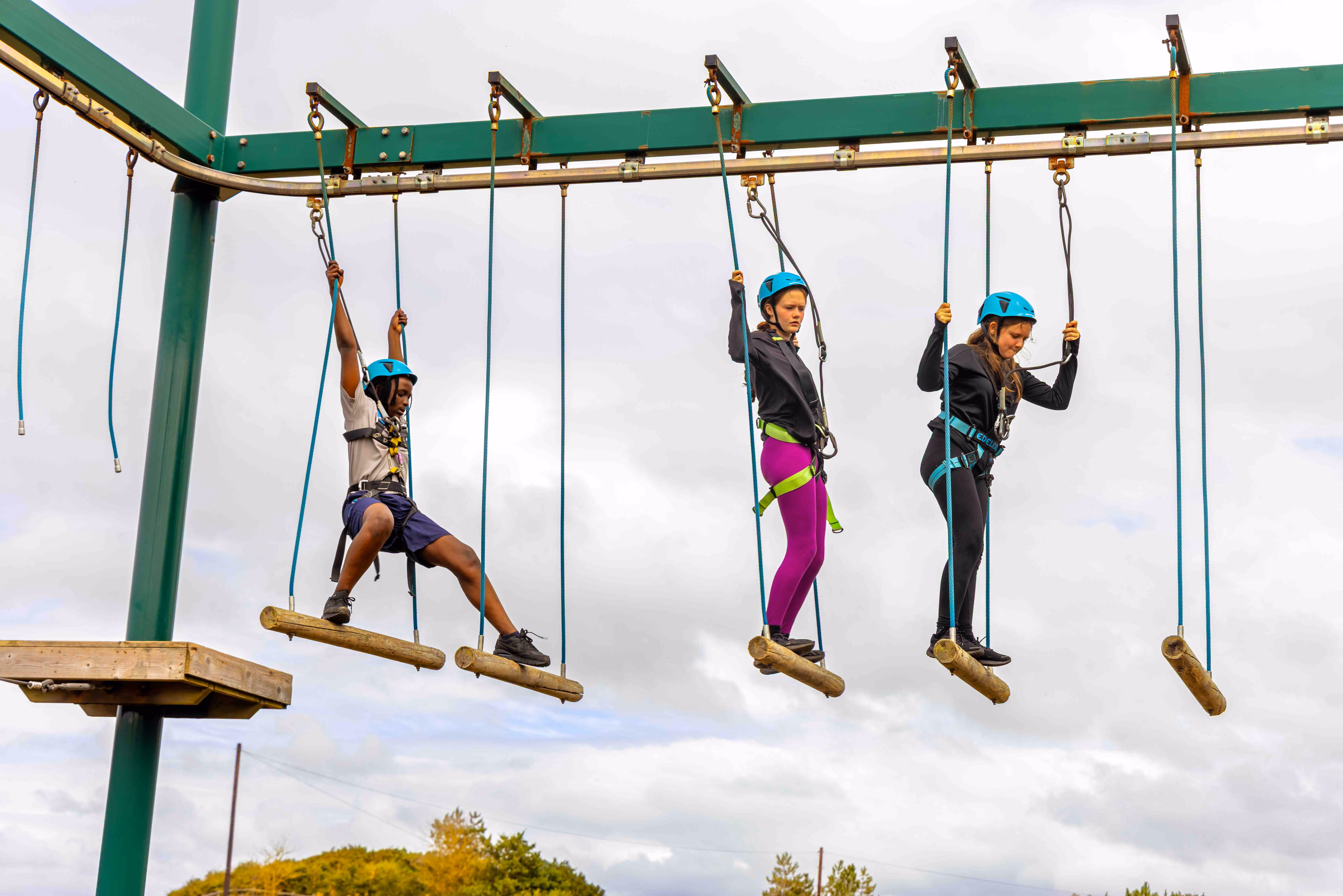 Three children wearing helmets and safety harnesses crossing suspended wooden log bridges at an outdoor ropes course.