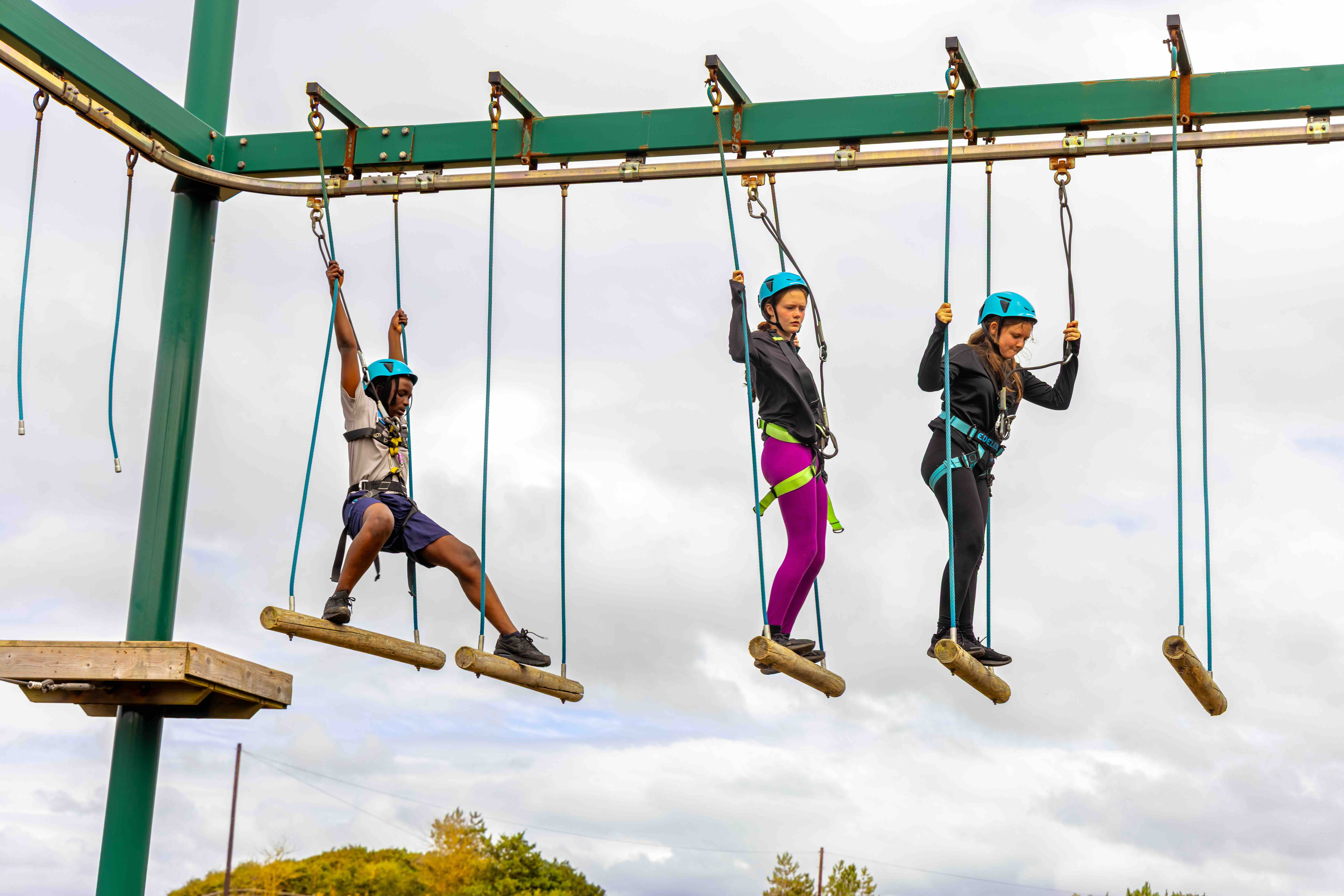Three children wearing helmets and safety harnesses crossing suspended wooden log bridges at an outdoor ropes course.