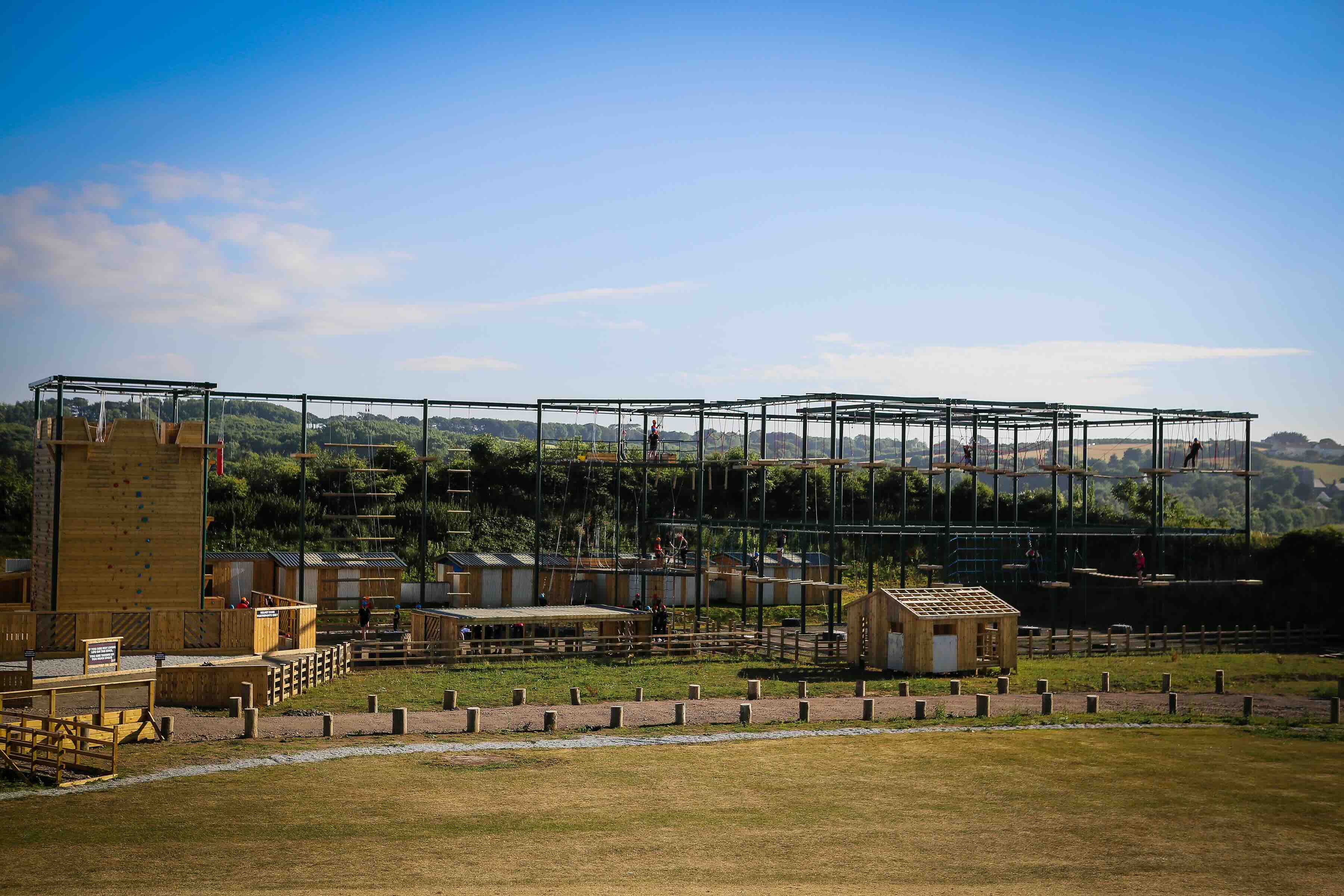 Outdoor ropes course with climbing wall, suspended platforms, and wooden cabins under a clear blue sky.