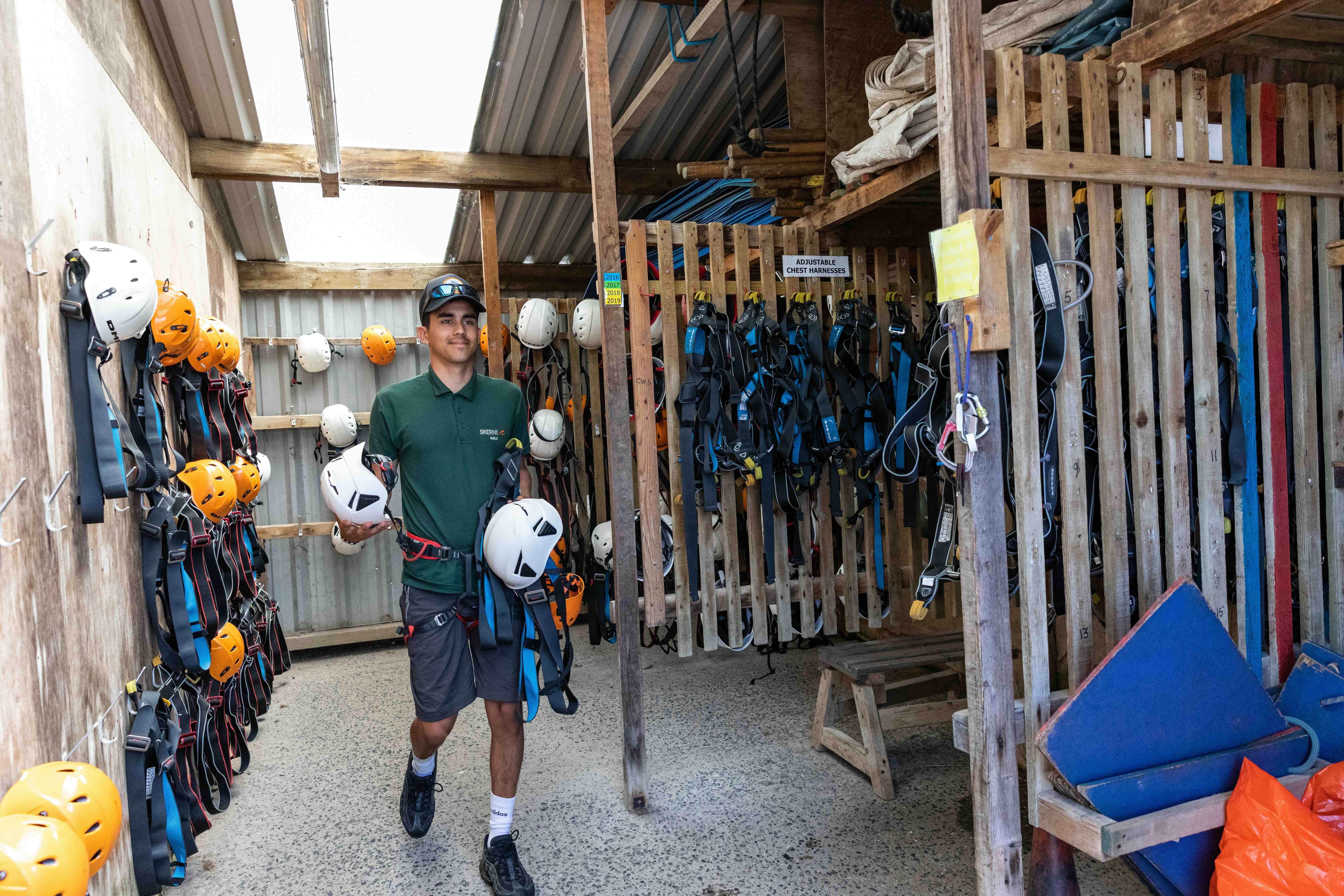 Man wearing a green polo and shorts holding safety helmets and harnesses in a wooden storage area filled with hanging helmets and adjustable chest harnesses.