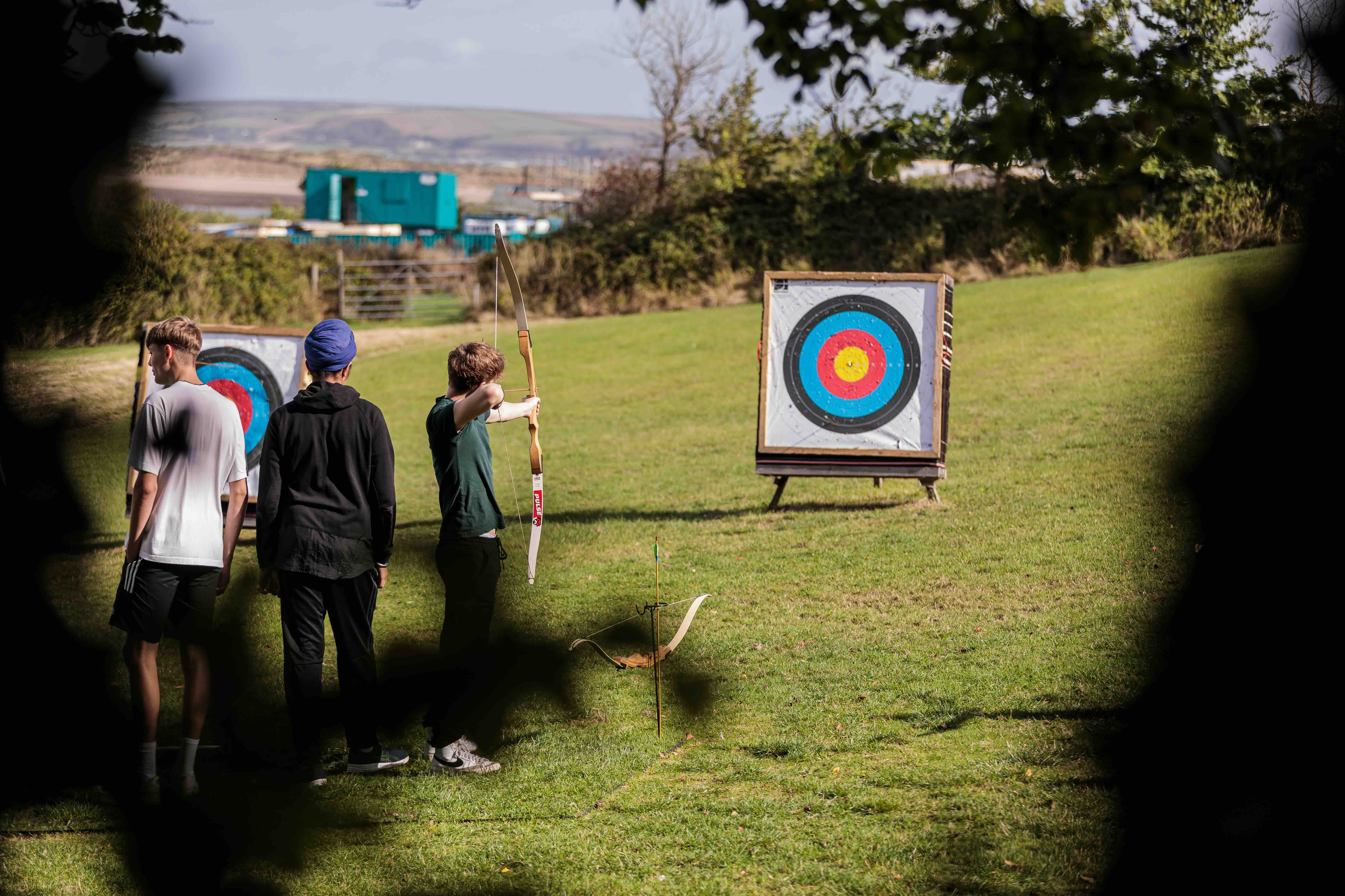 Three young people standing on grass with one aiming a bow and arrow at an archery target in an outdoor setting.