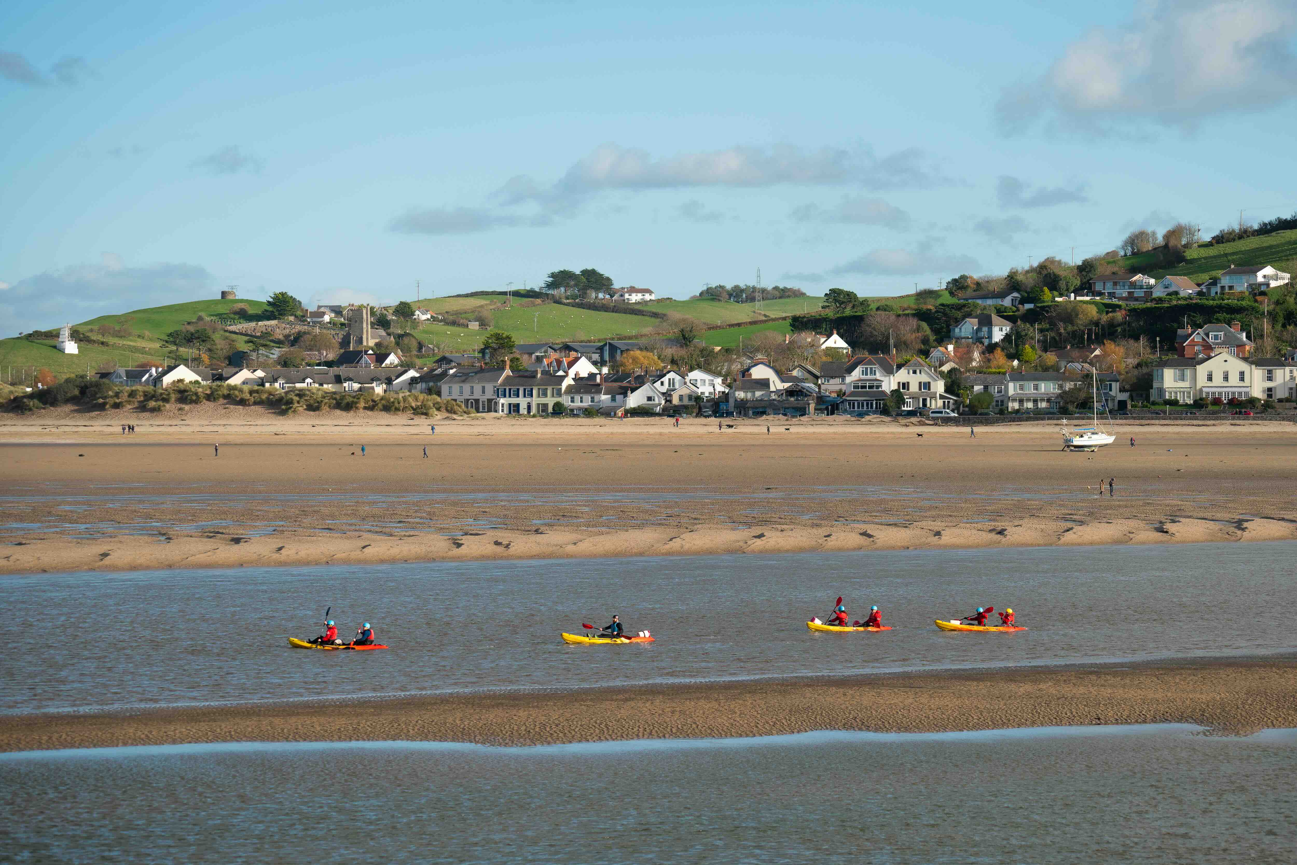 Group of people kayaking on a calm river near a sandy beach with houses and green hills in the background under a blue sky.