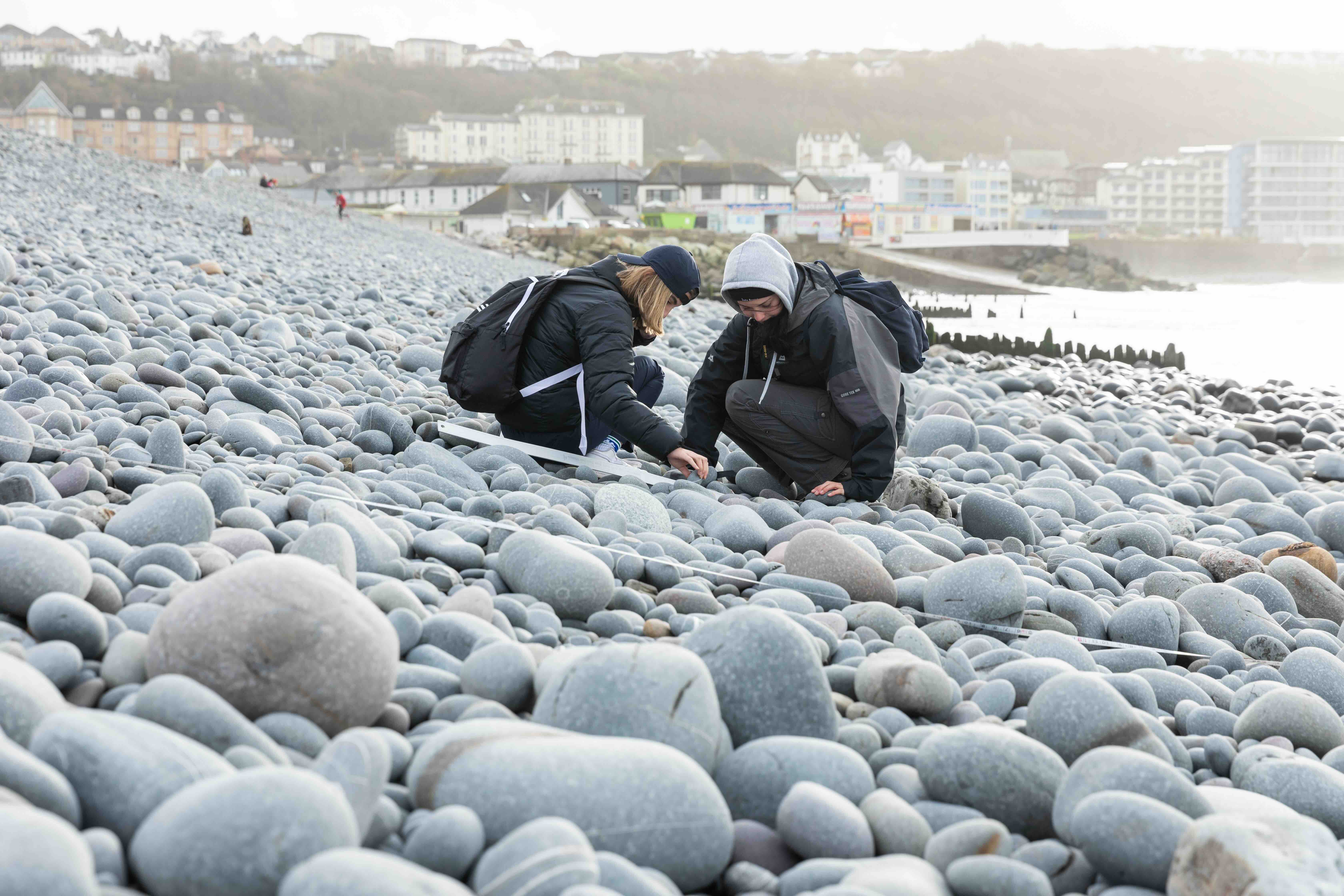 Two people in outdoor jackets examining rocks on a pebble beach near a coastal town.