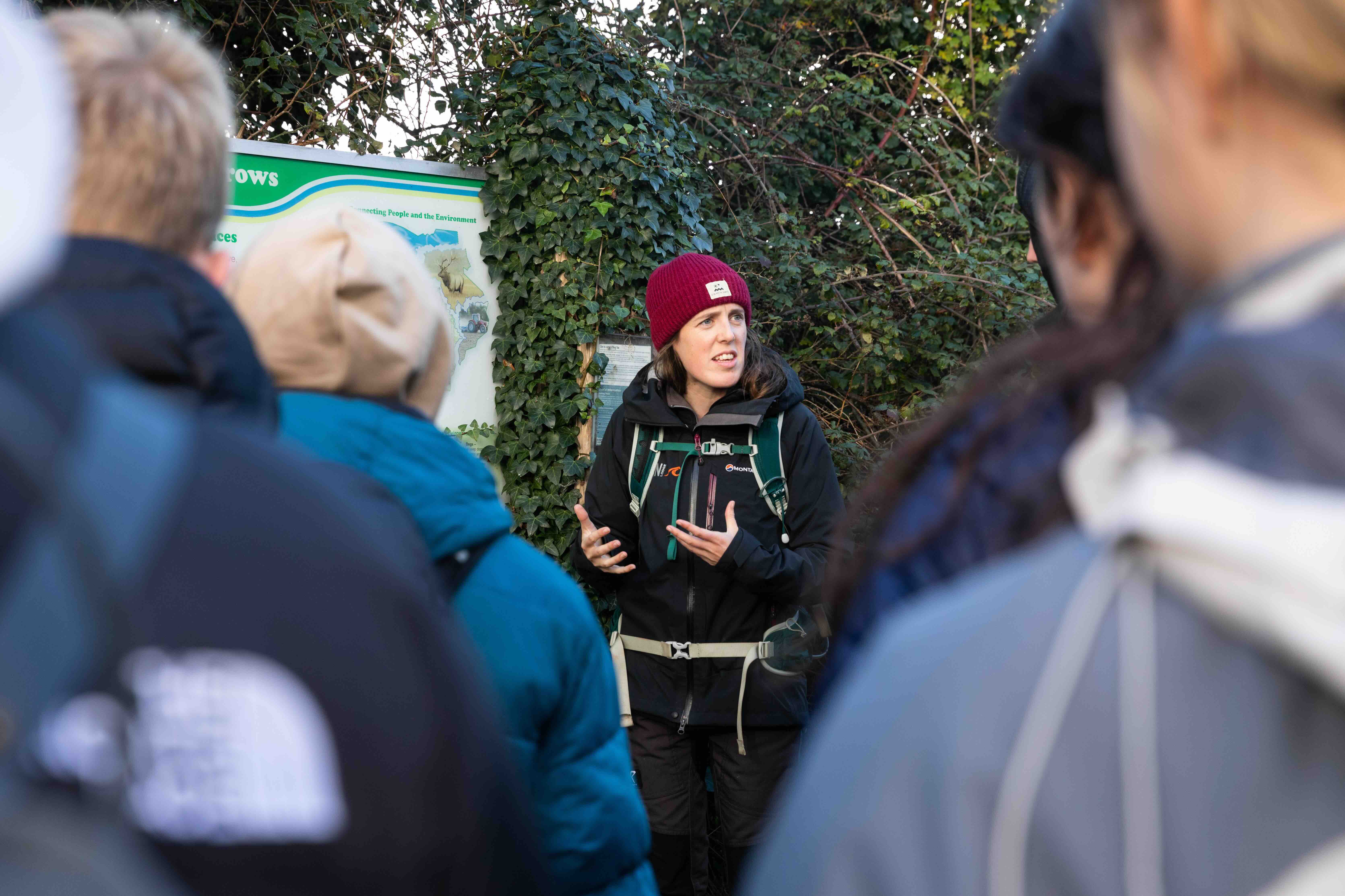 Woman in outdoor gear and a red beanie explaining information to a group in front of a green sign and ivy-covered wall.