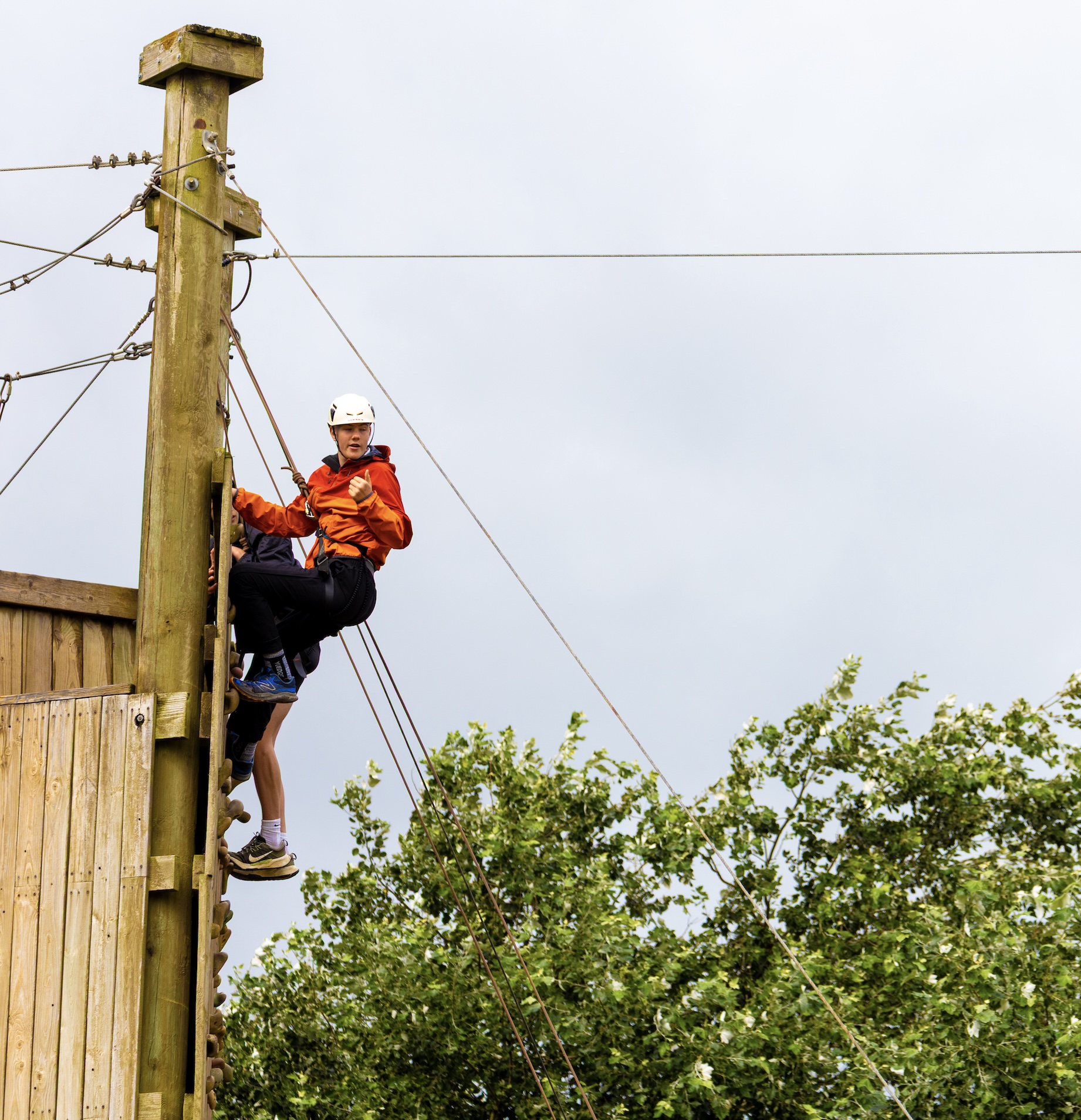 Person in an orange jacket and white helmet climbing a wooden structure secured with ropes outdoors.