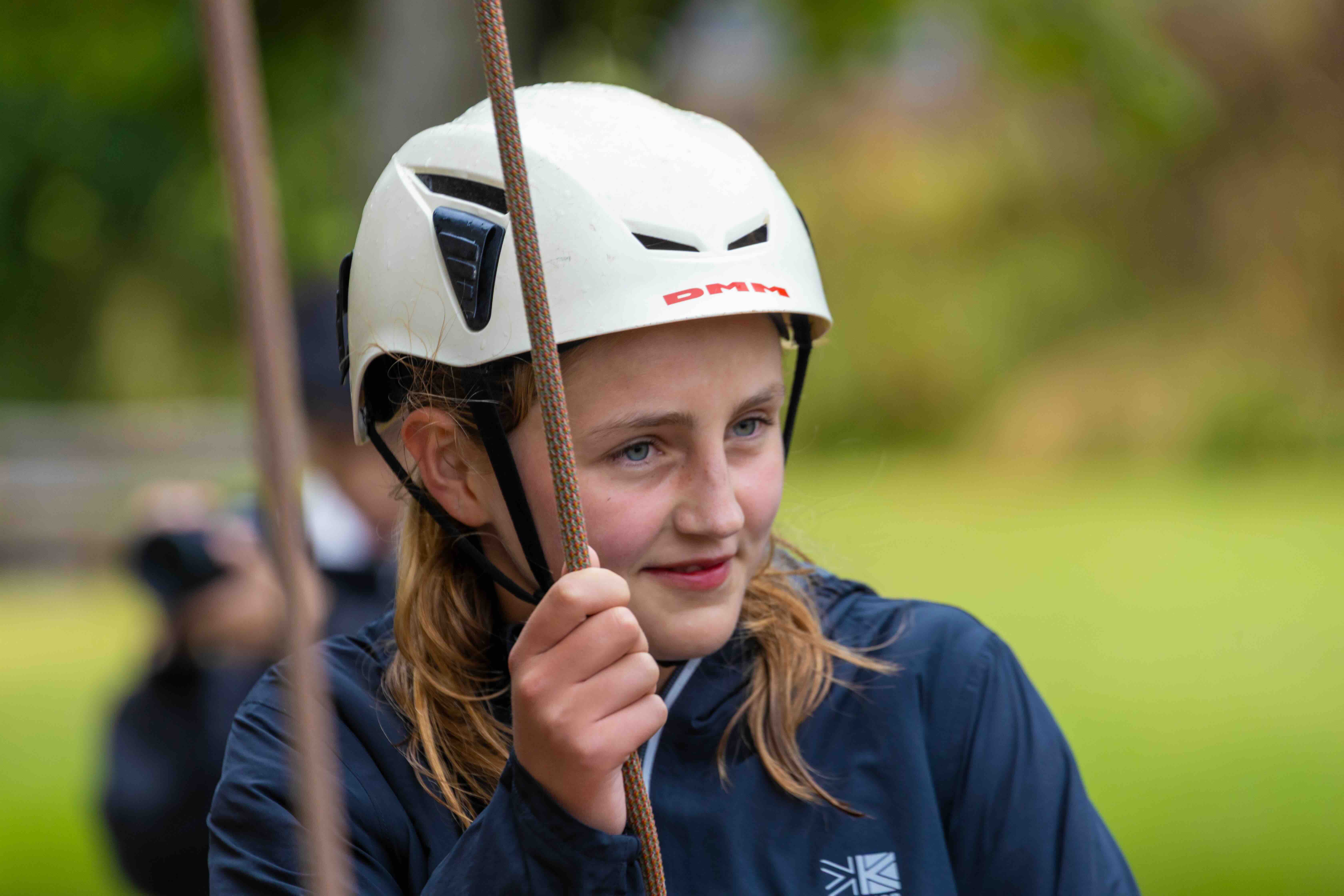 Young girl wearing a white helmet and navy jacket holding a climbing rope outdoors.