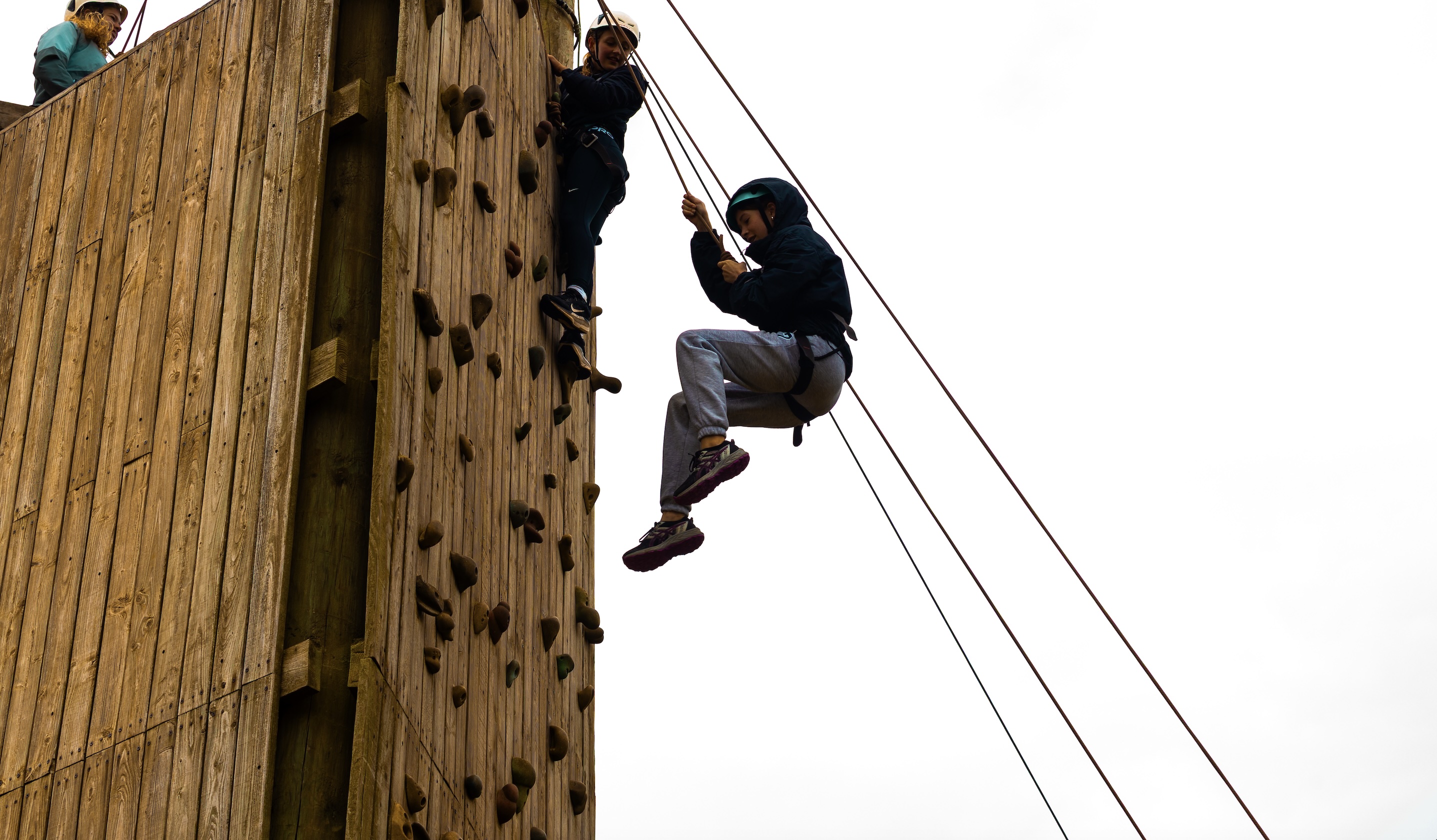 Two people climbing and rappelling down a tall wooden climbing wall with safety harnesses and helmets.