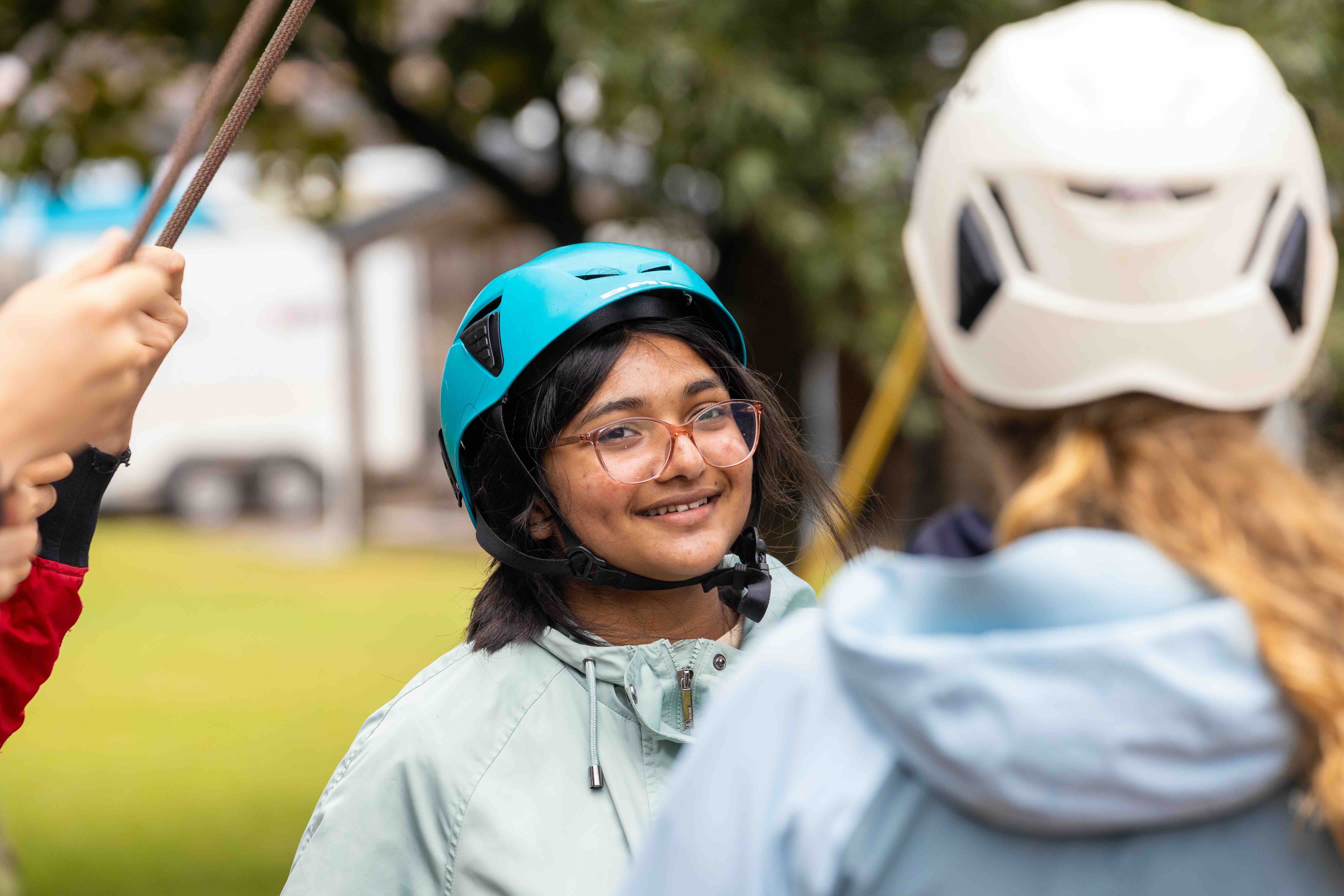 Smiling young woman wearing a blue helmet and glasses speaking outdoors with another person in a white helmet.