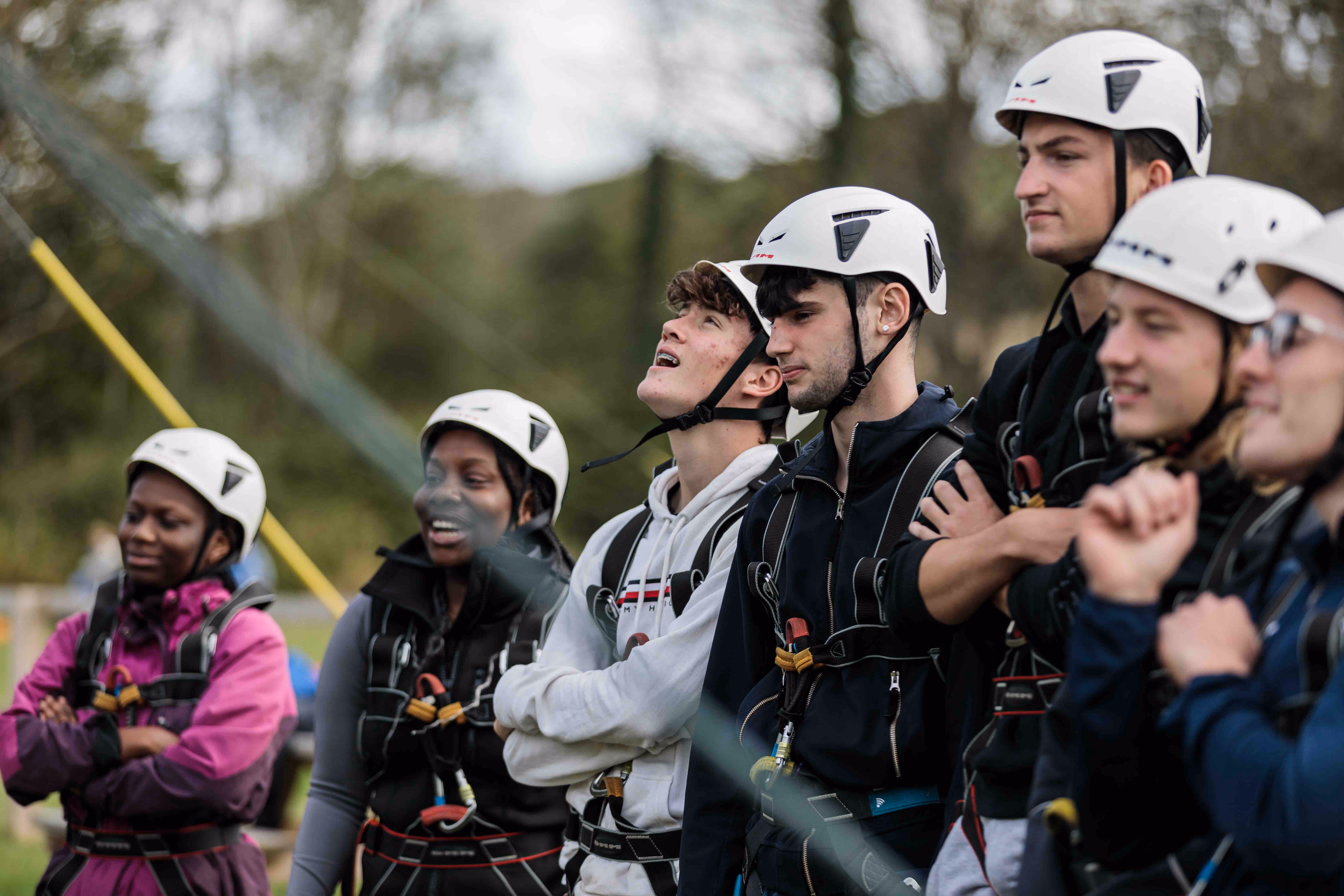 Group of young people wearing helmets and harnesses outdoors, some smiling and looking upwards.