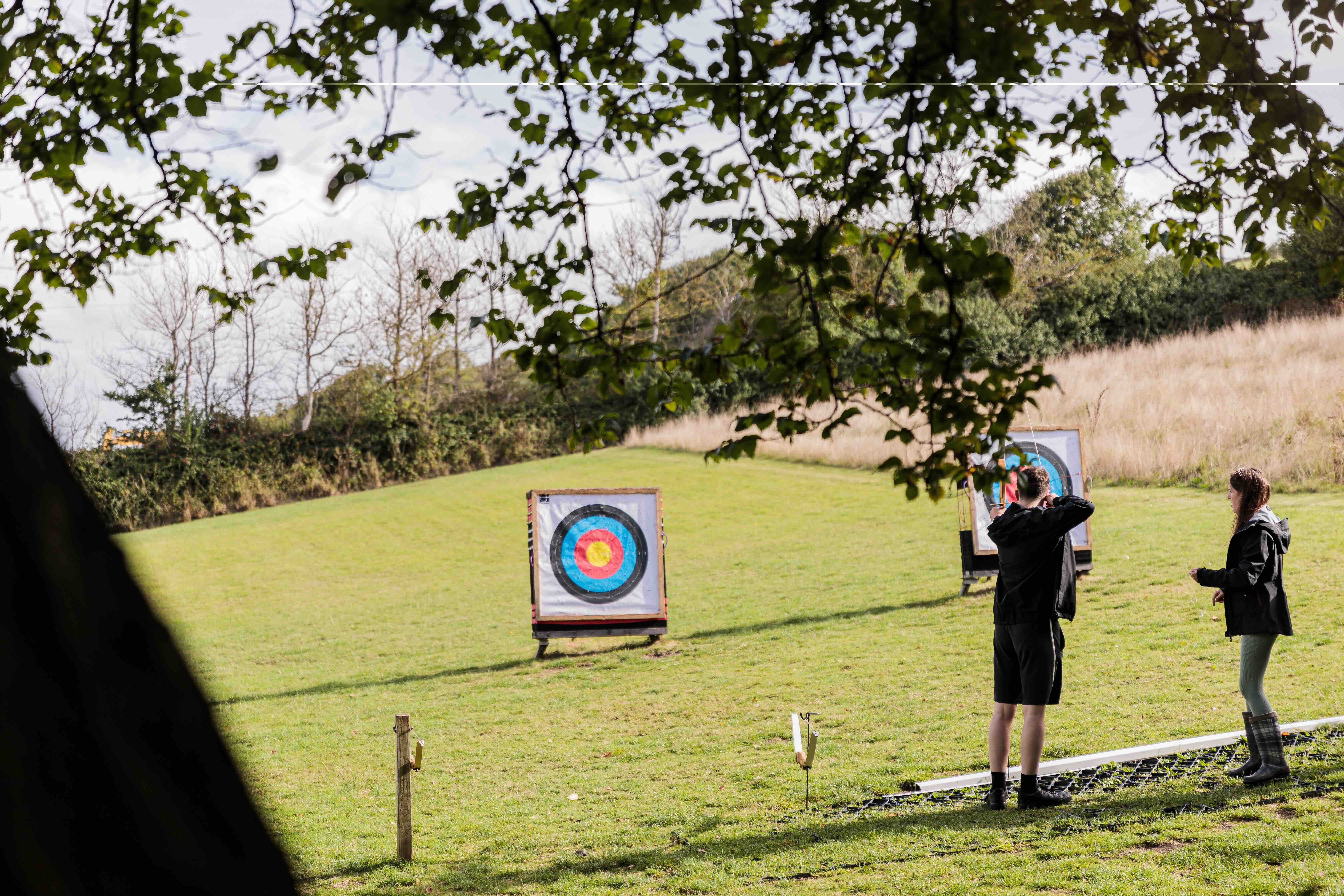 Two people standing on grass aiming at archery targets on a sunny field with trees in the background.