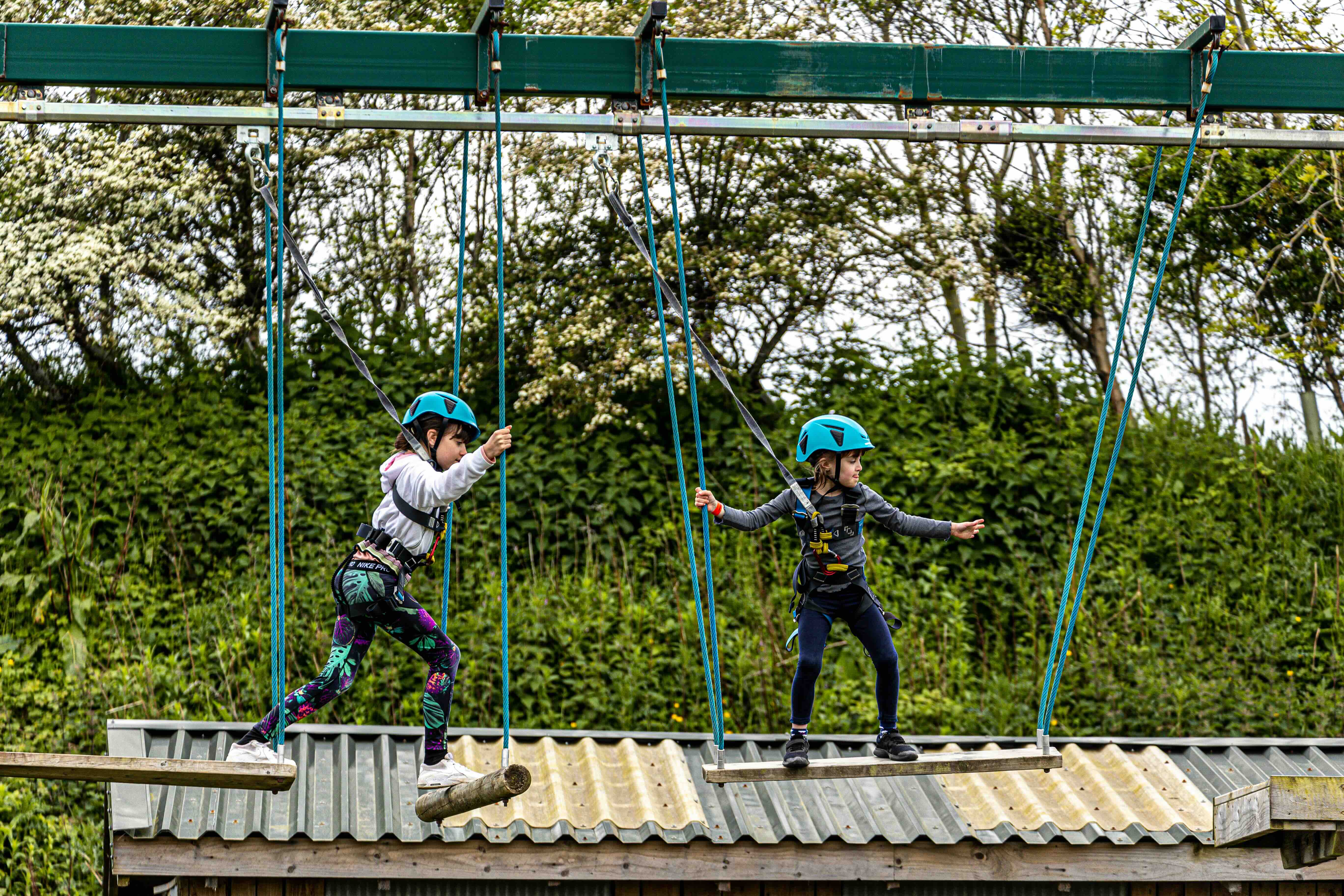 Two children wearing blue helmets and safety harnesses navigating a high ropes course obstacle suspended above a corrugated metal roof with greenery in the background.