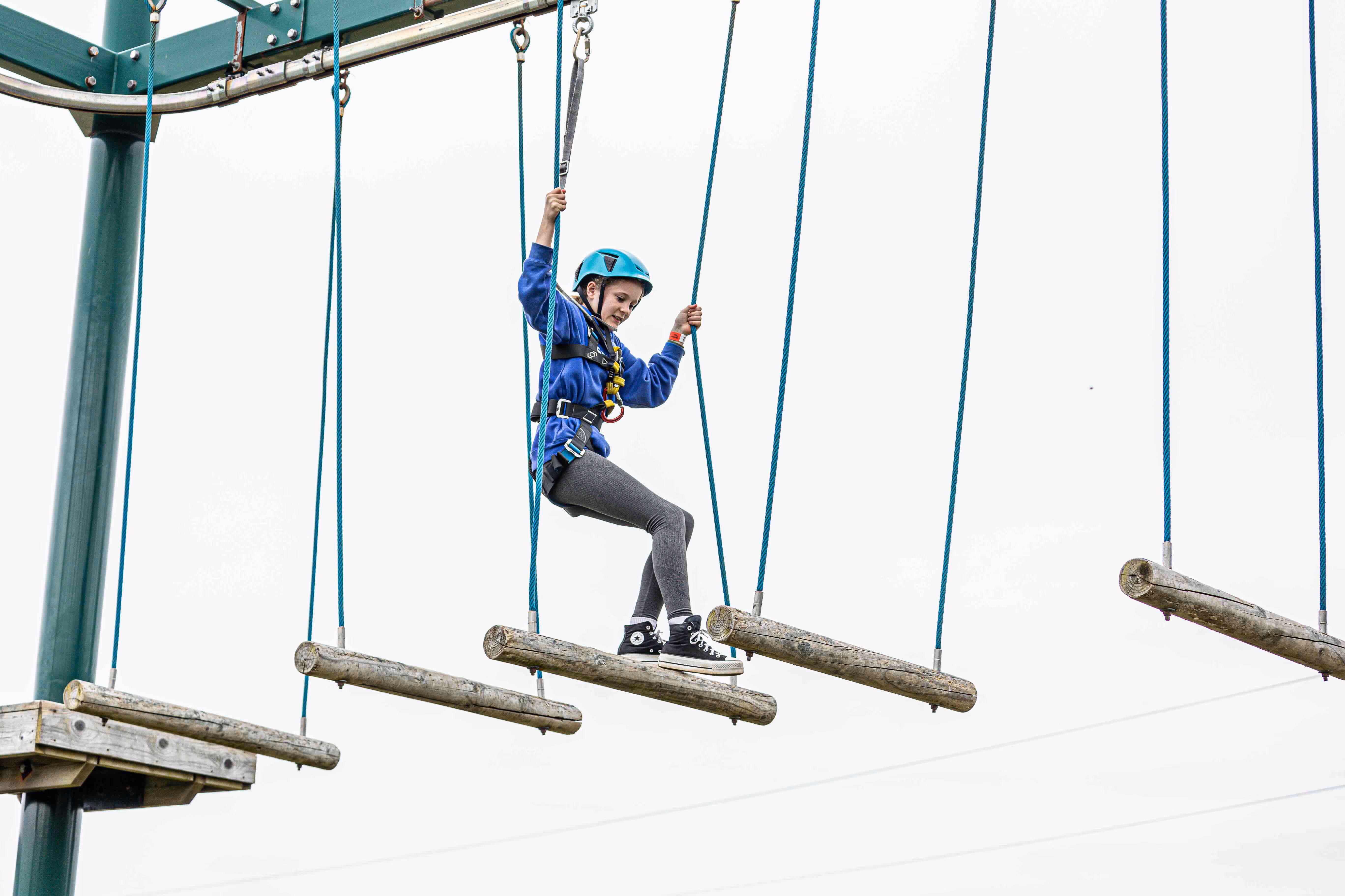 Girl wearing a blue helmet and harness navigating suspended wooden logs on a high ropes course.
