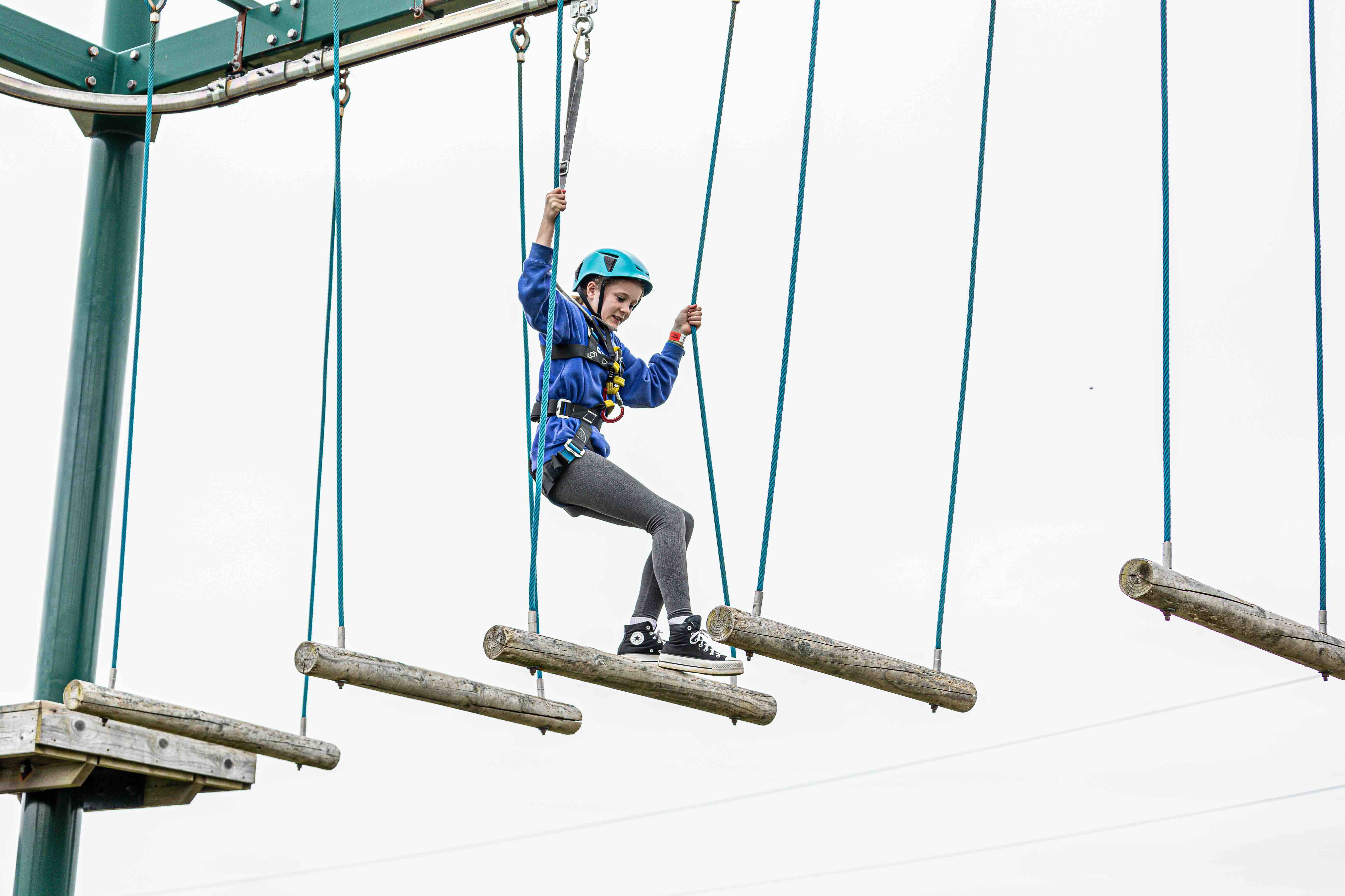Girl wearing a blue helmet and harness navigating suspended wooden logs on a high ropes course.