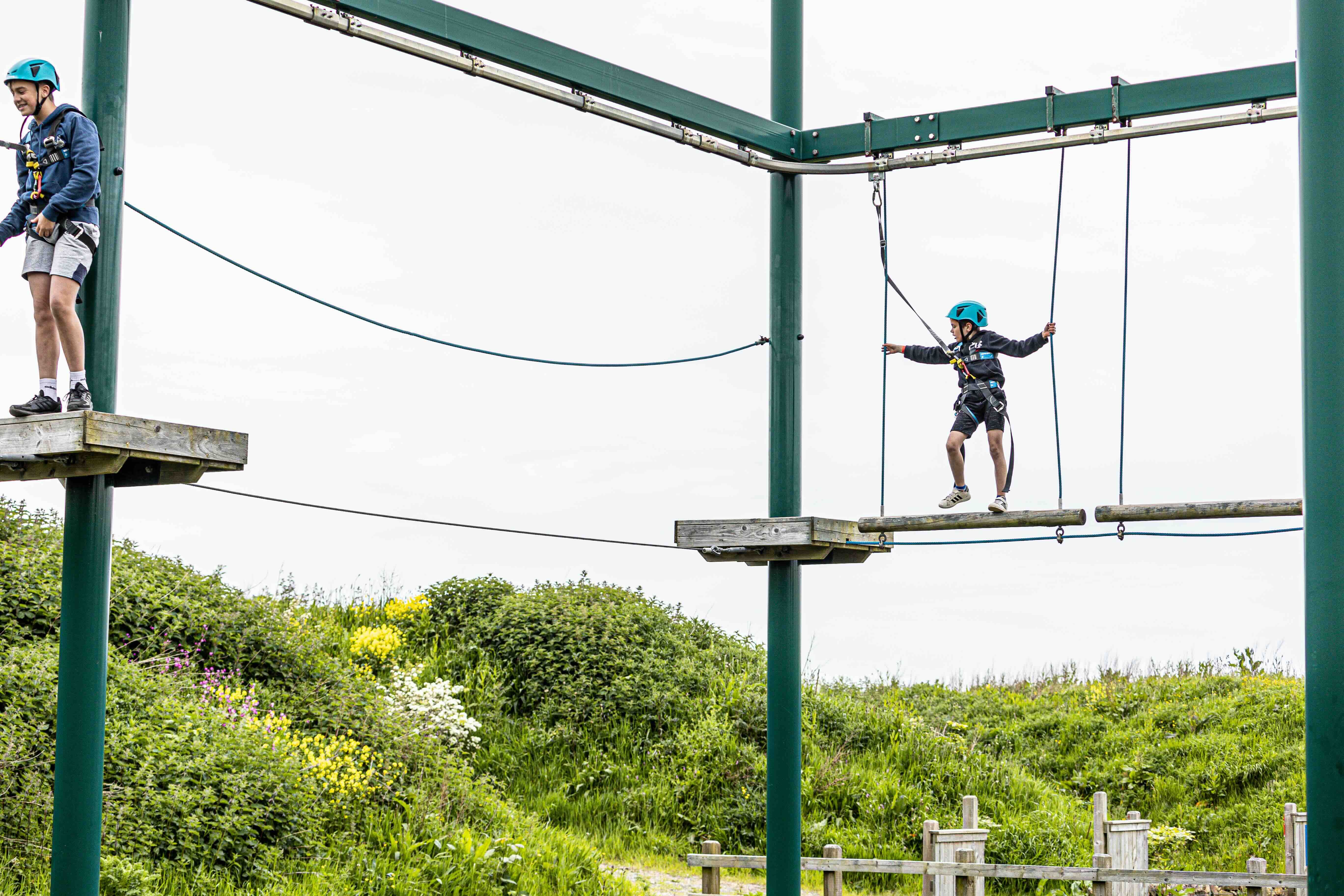Two boys wearing helmets and harnesses participating in a high ropes course outdoors.