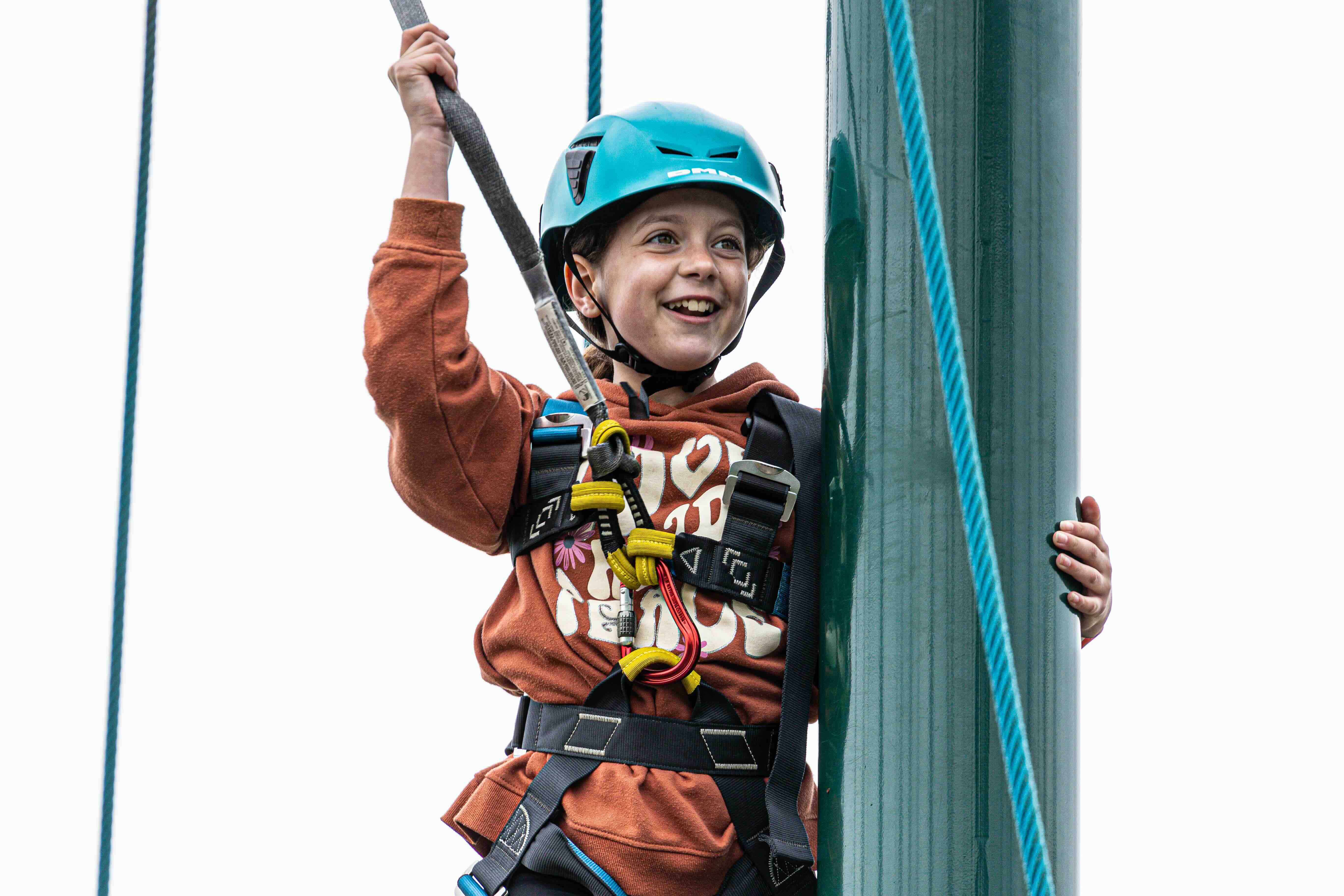 Smiling child wearing a blue helmet and safety harness climbing a green pole in a high ropes course.