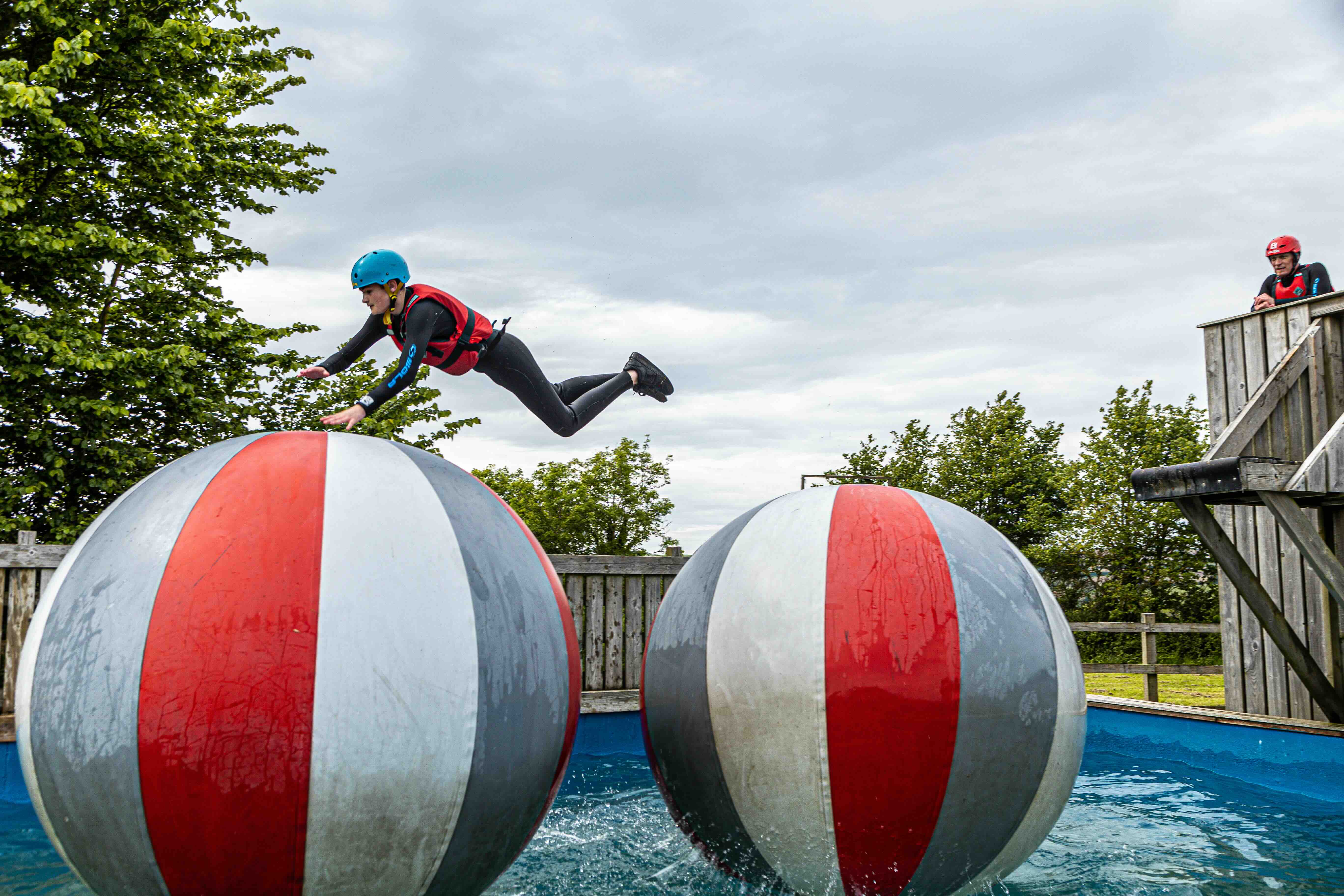 Person in a blue helmet and red vest jumping mid-air between two large red, white, and grey inflated balls over water.
