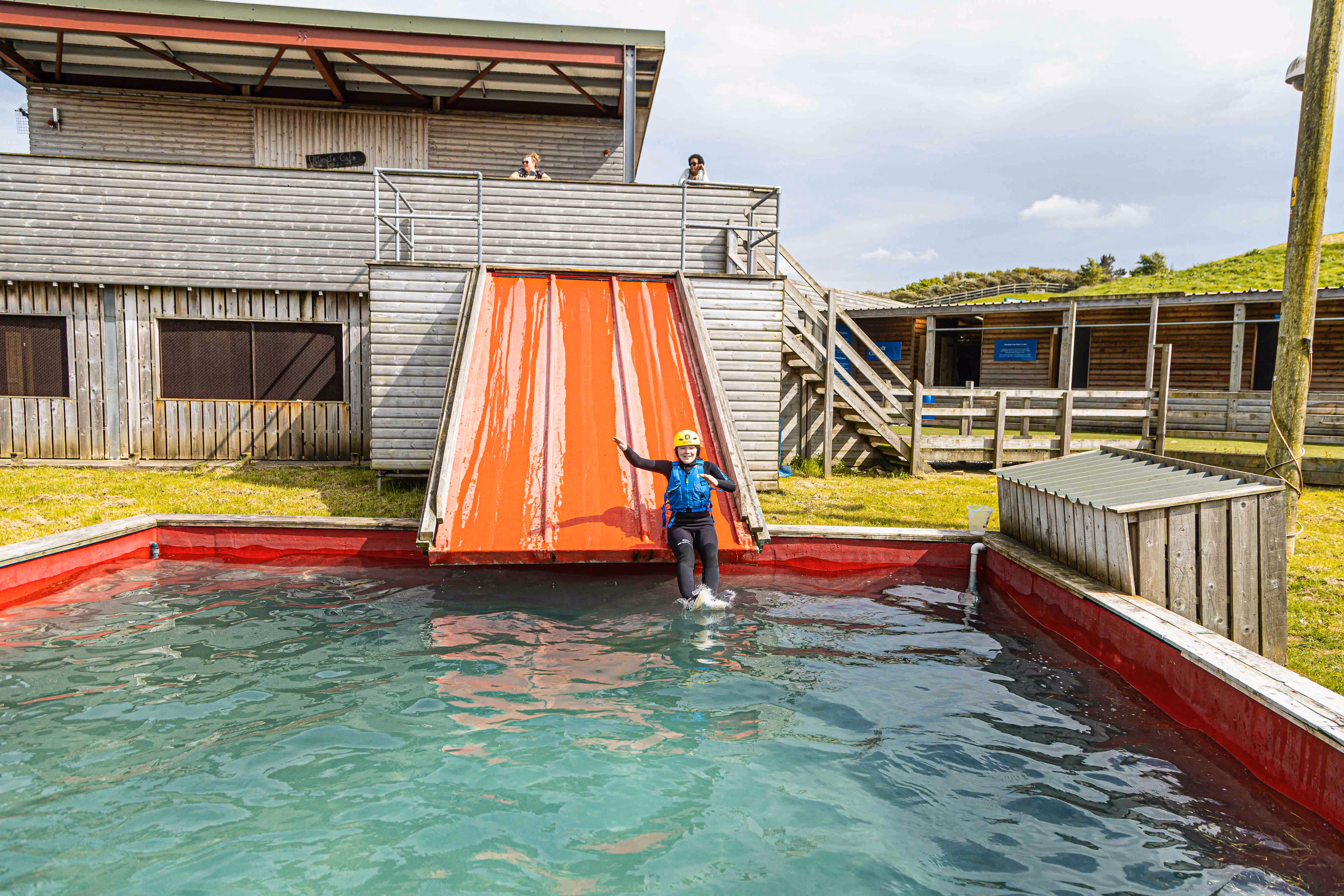 Person wearing a yellow helmet and safety vest sliding down a wet orange ramp into a water pool outside a wooden building.