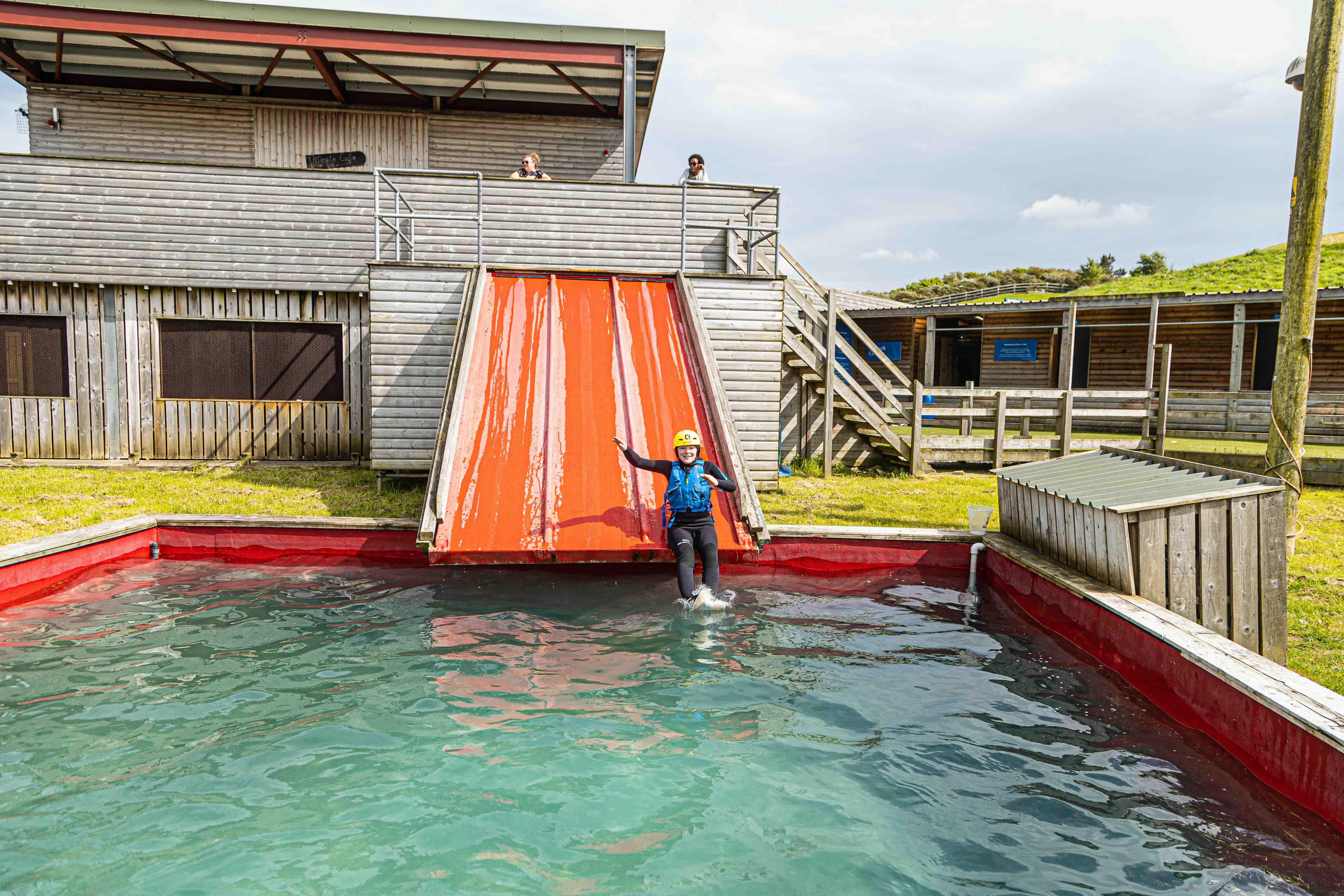 Person wearing a yellow helmet and safety vest sliding down a wet orange ramp into a water pool outside a wooden building.