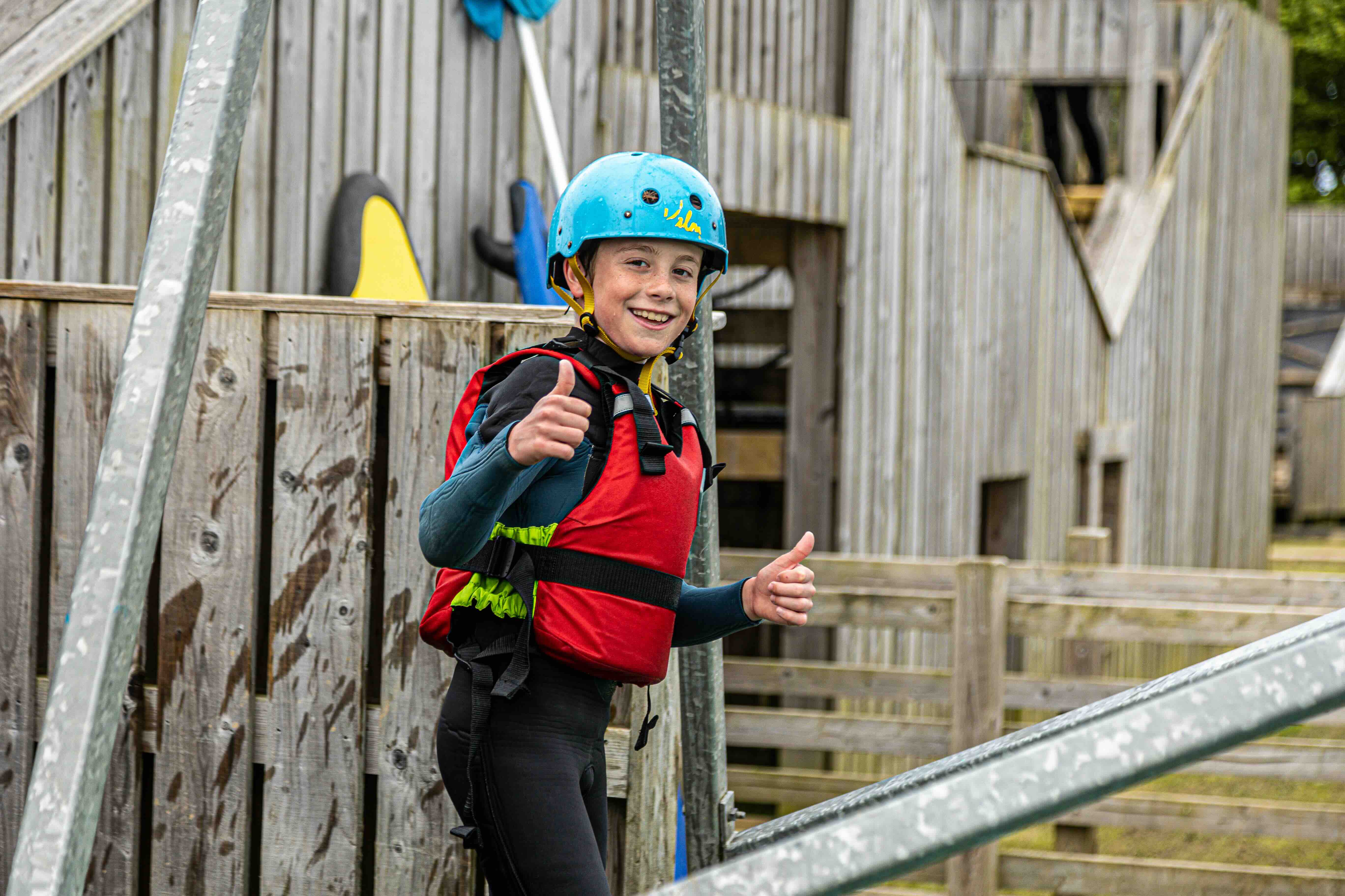 Smiling boy wearing a blue helmet and red life jacket giving two thumbs up at an outdoor obstacle course.