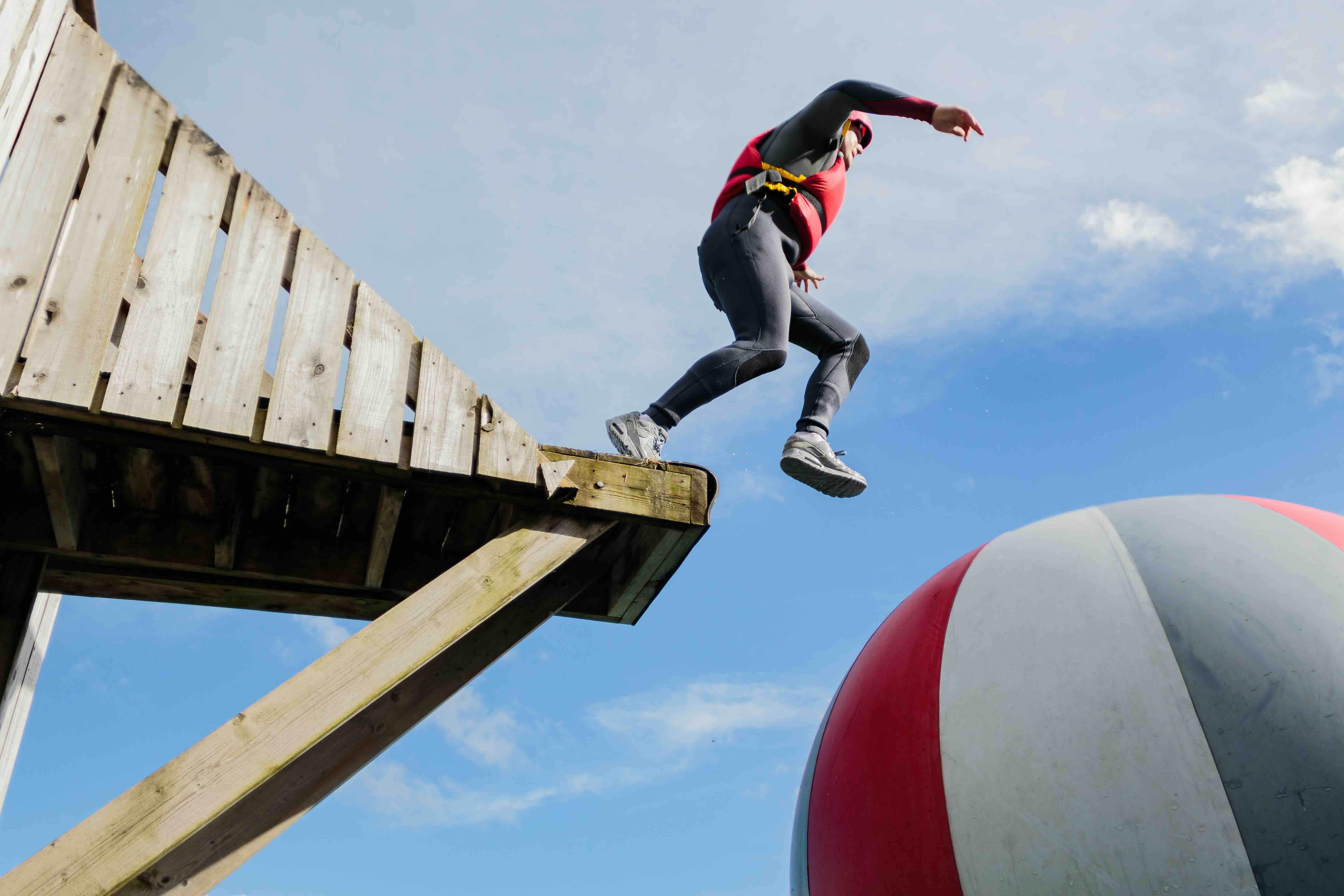 Person in a wetsuit and helmet jumping from a wooden platform onto a large inflatable ball outdoors.