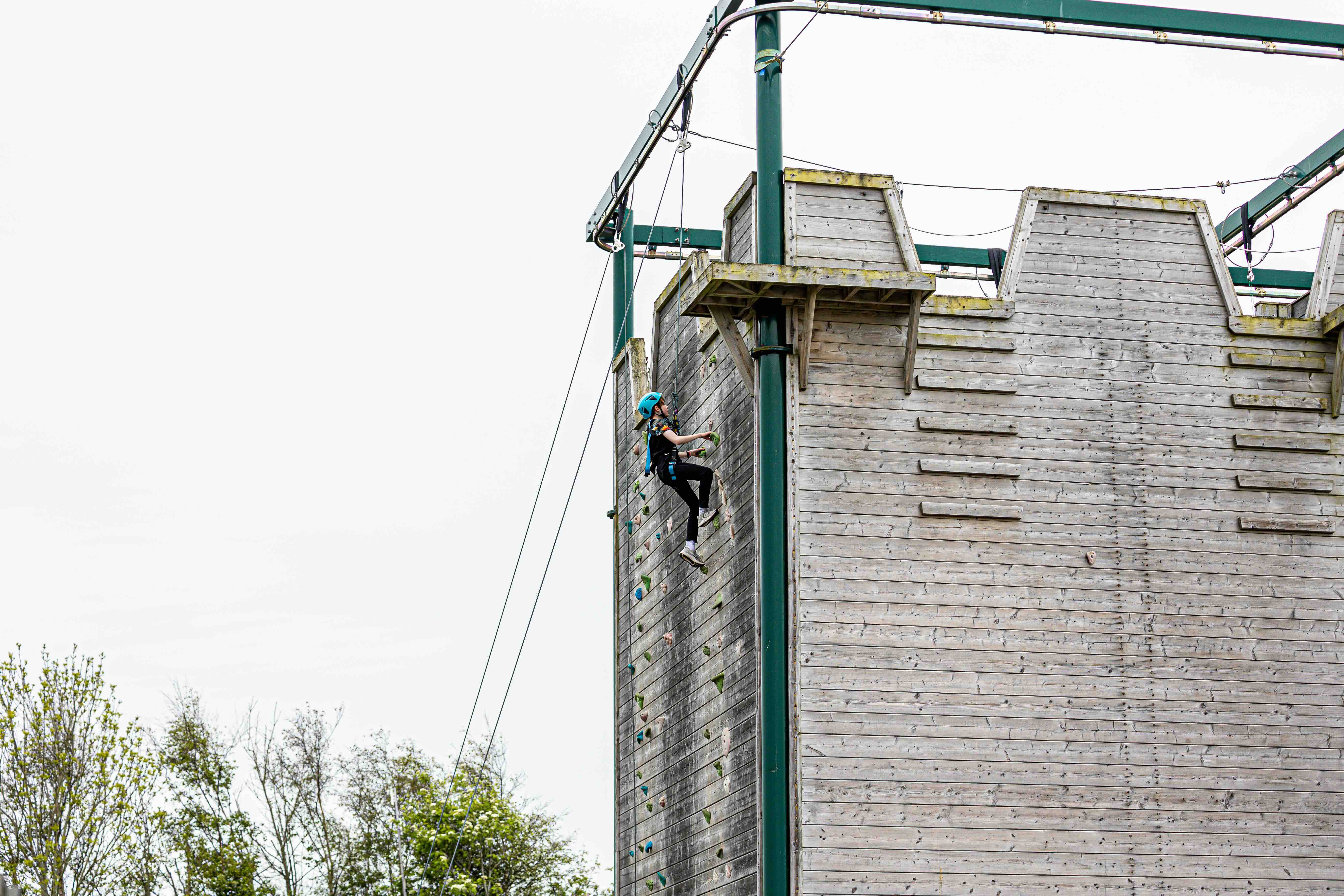 Person wearing a helmet climbing a tall outdoor wooden climbing wall with harness and ropes.