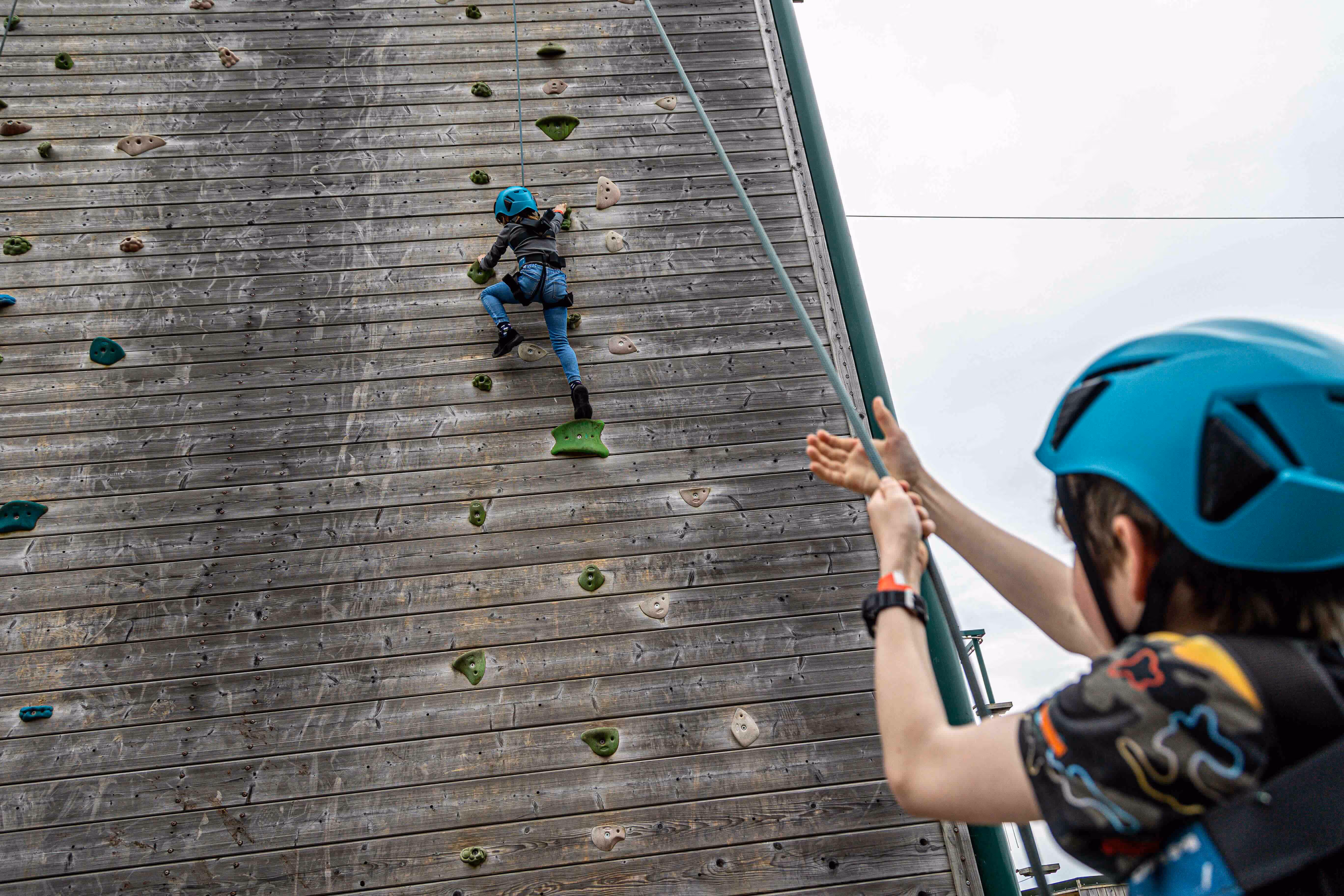 Child wearing a blue helmet climbing a wooden climbing wall while another child in a helmet holds the safety rope.