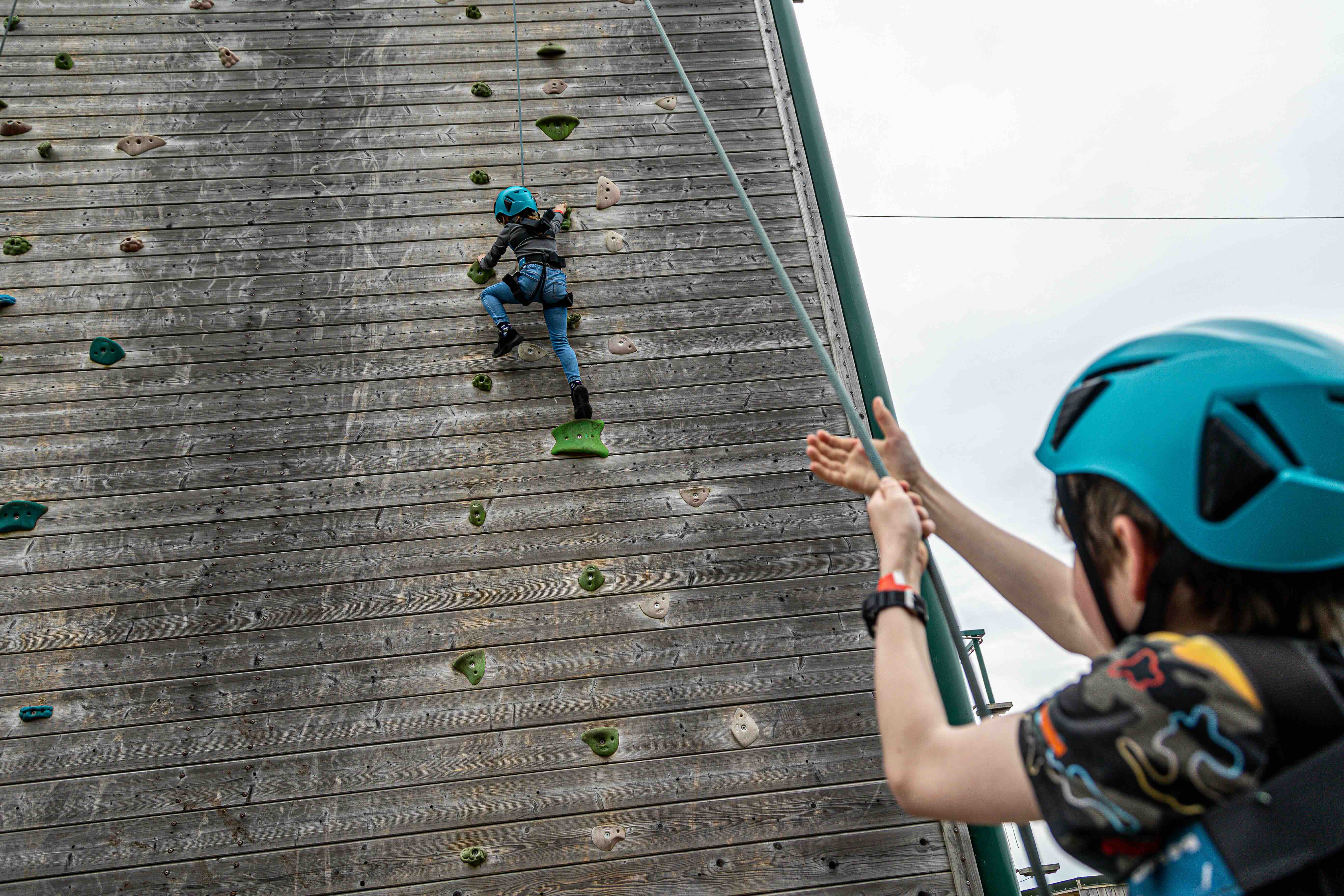 Child wearing a blue helmet climbing a wooden climbing wall while another child in a helmet holds the safety rope.