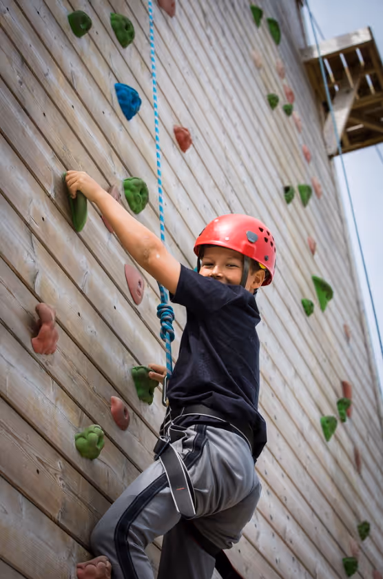 Child wearing a red helmet climbing an outdoor wooden rock climbing wall with colourful holds.