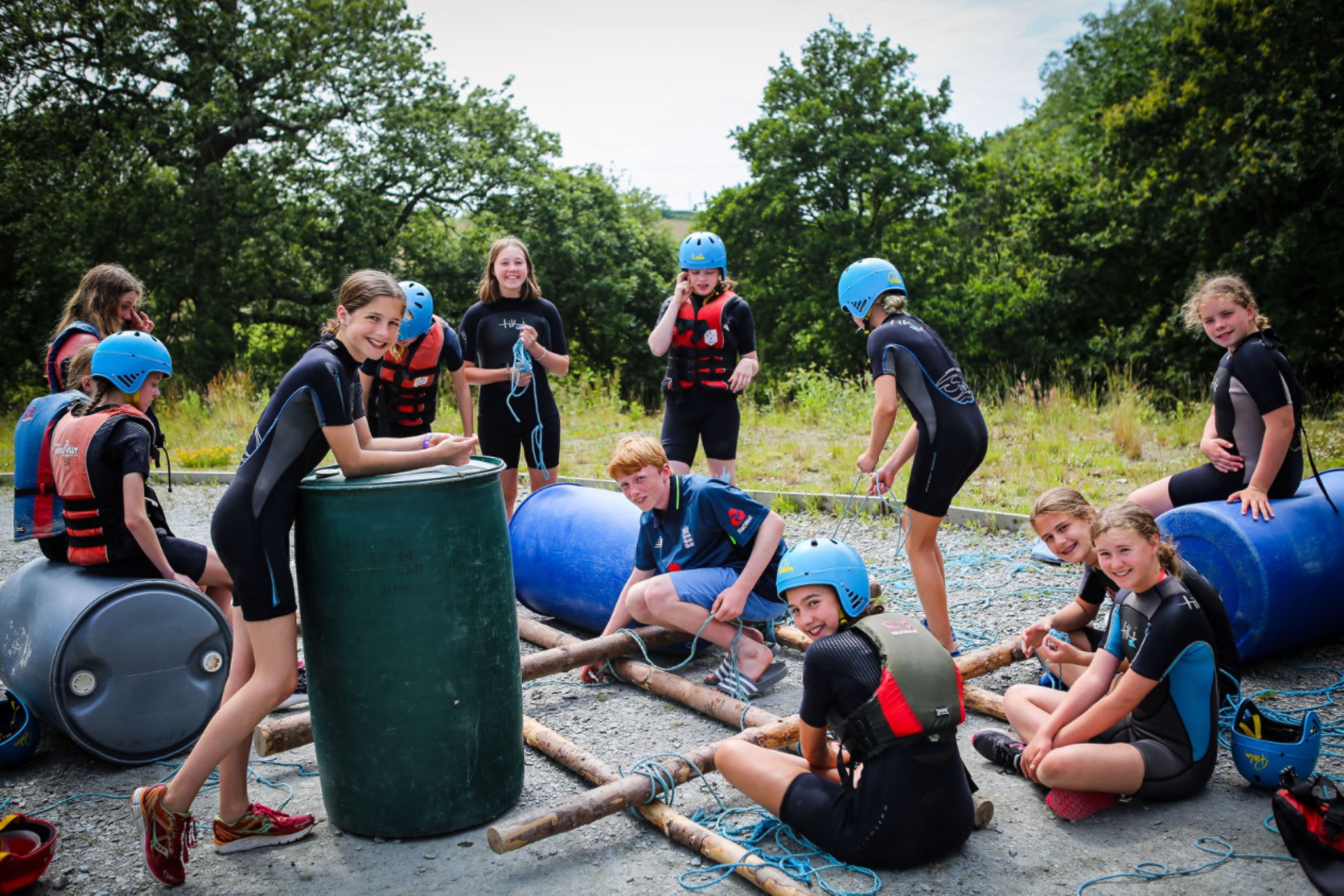 Group of children outdoors building a raft using logs, barrels, ropes, wearing wetsuits, helmets and life jackets.
