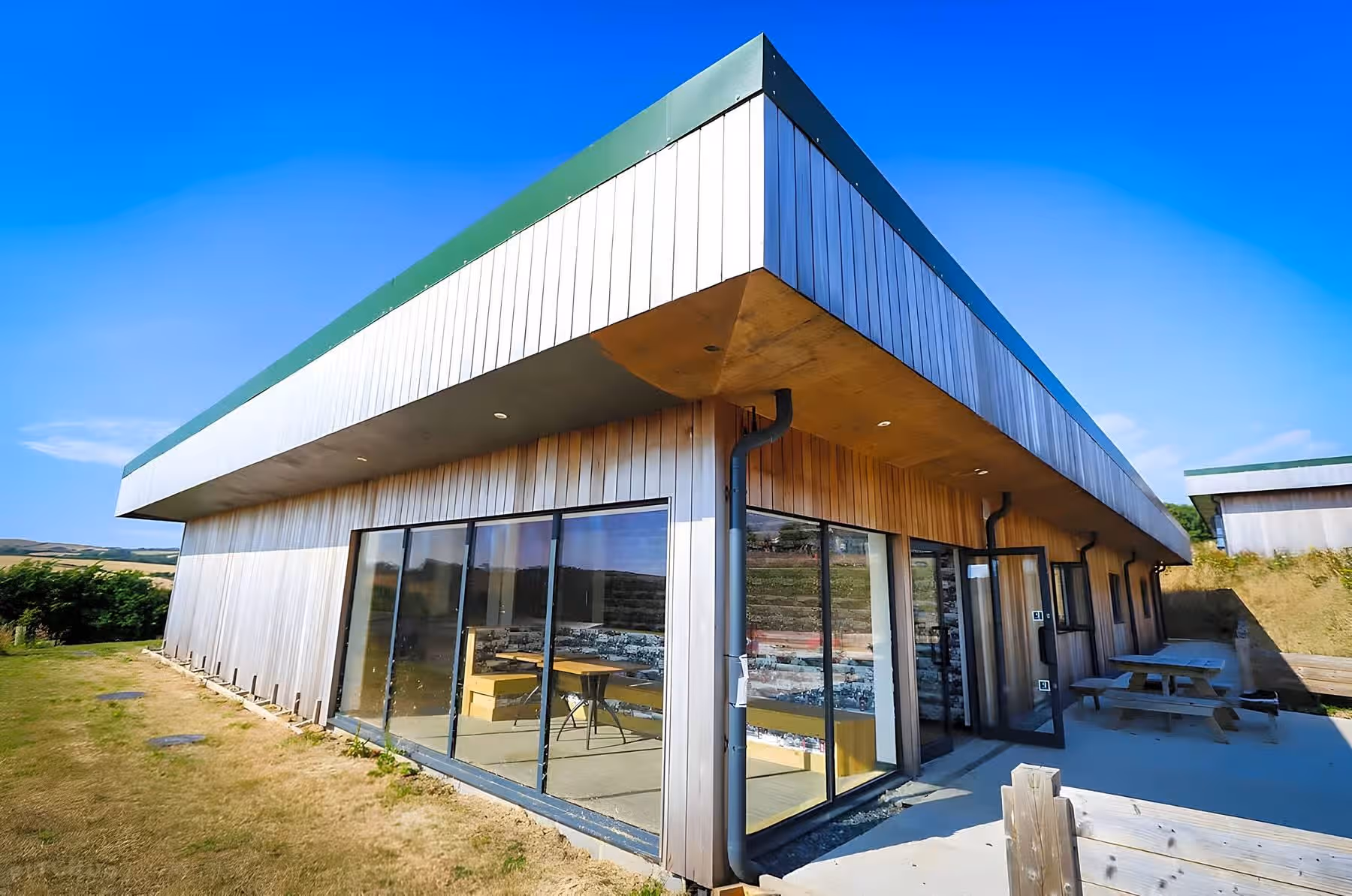 Modern wooden building with large glass windows and picnic tables outside on a sunny day.