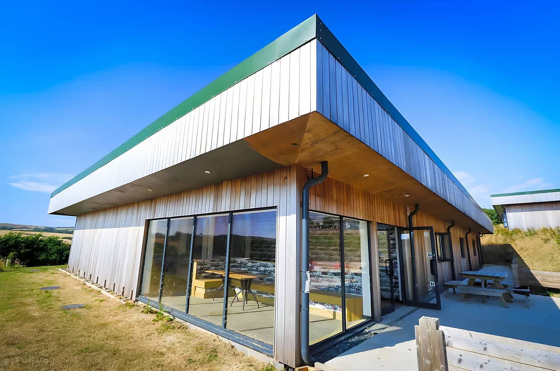 Modern wooden building with large glass windows and picnic tables outside on a sunny day.
