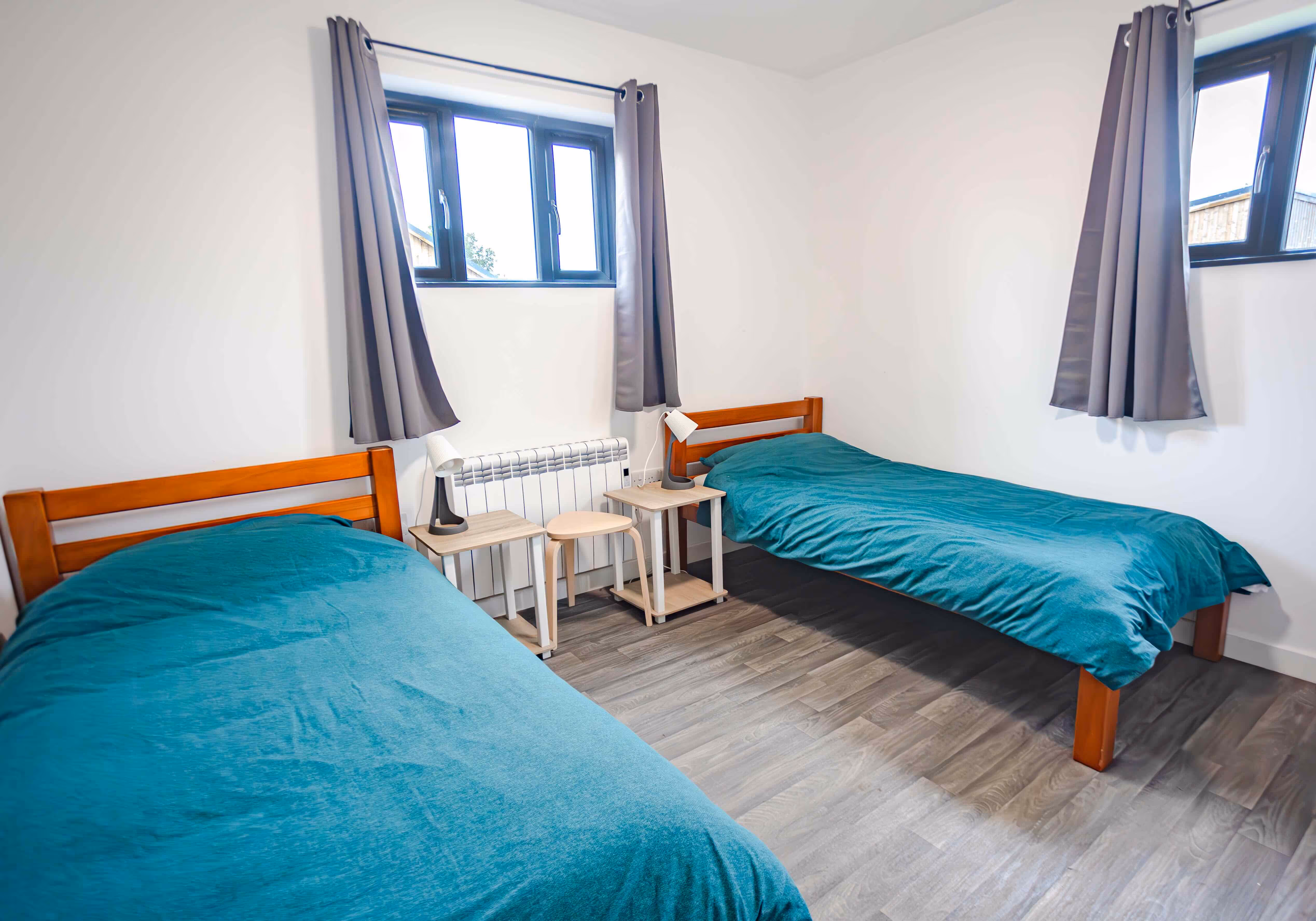 Minimalist bedroom with two single wooden beds covered in teal bedding, two bedside tables with matching lamps, grey curtains, and wooden flooring.