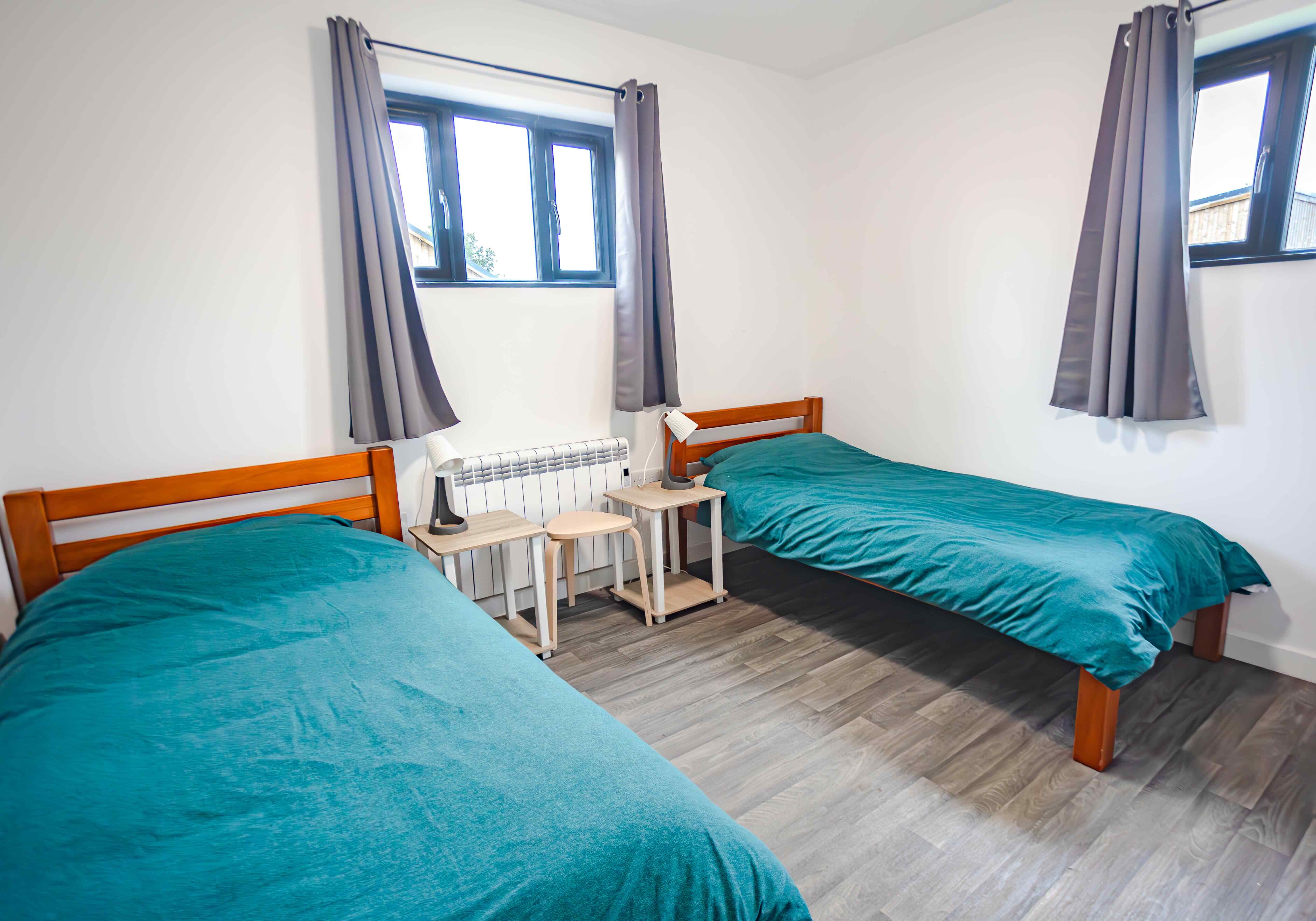 Minimalist bedroom with two single wooden beds covered in teal bedding, two bedside tables with matching lamps, grey curtains, and wooden flooring.