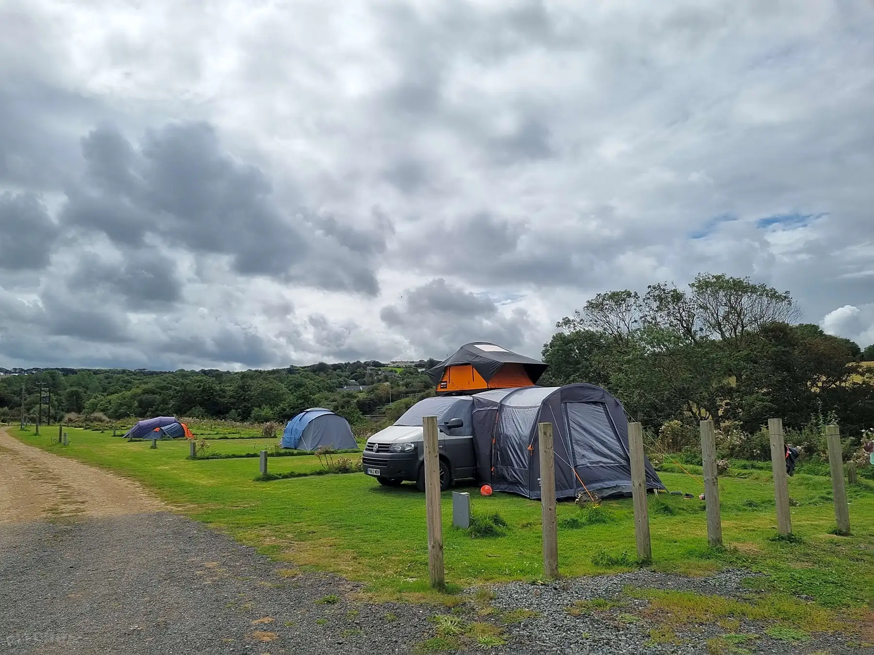 Campervan with roof tent and attached awning tent on a grassy campsite under a cloudy sky, with other tents in the background.