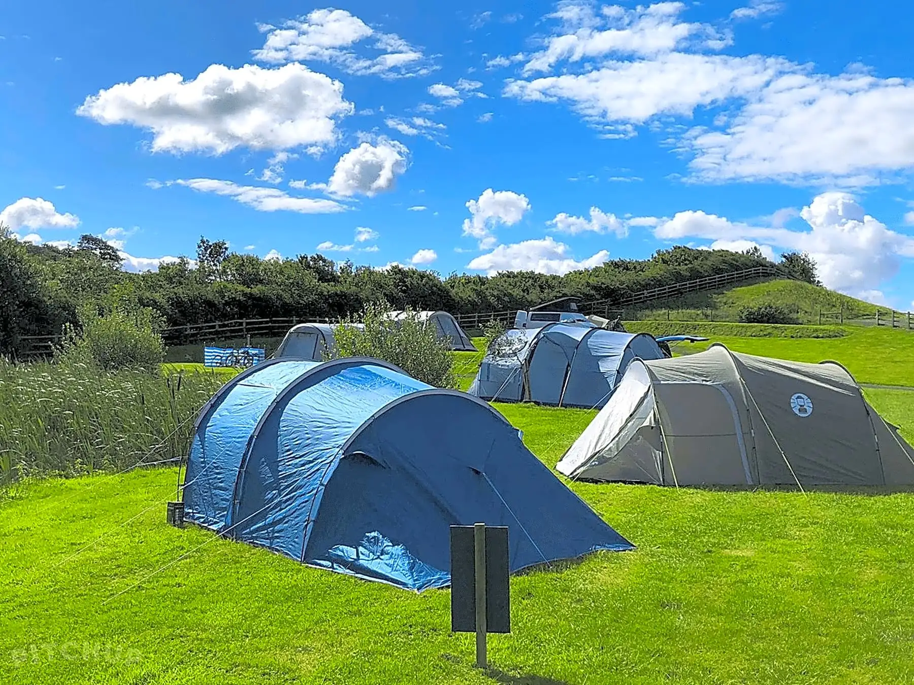 Blue and grey tents pitched on bright green grass under a blue sky with scattered white clouds.